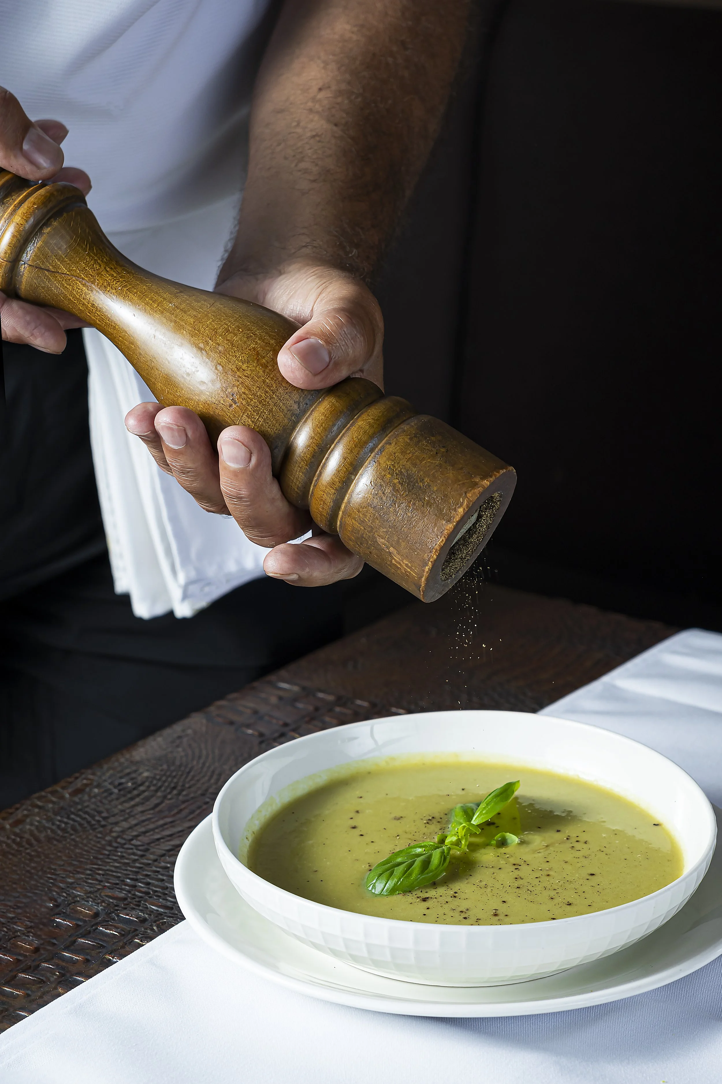 A bowl of green soup garnished with fresh basil leaves on a white plate, with a person holding a wooden pepper grinder over it.