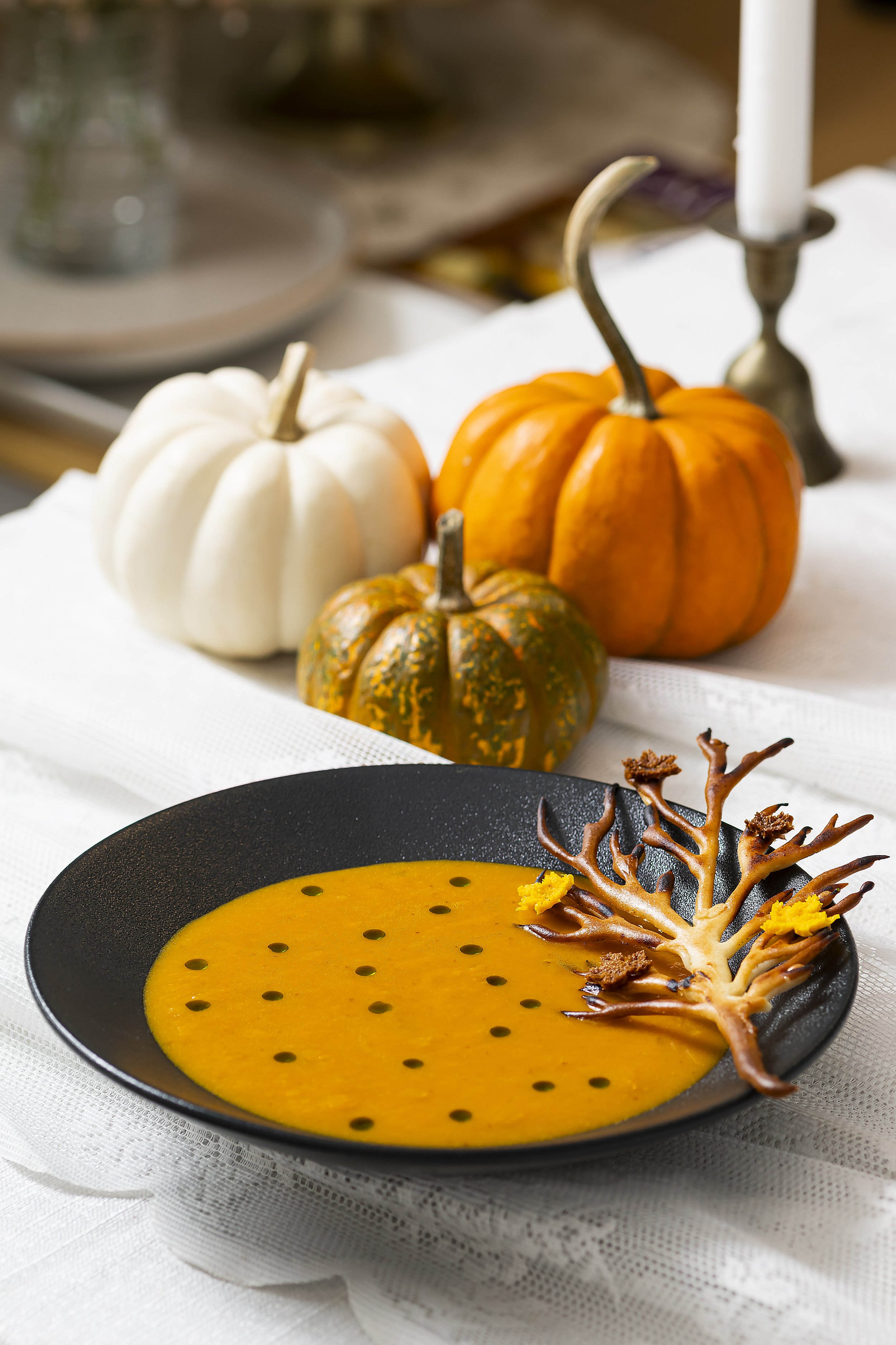 A black bowl of bright orange pumpkin soup garnished with a decorative edible branch and yellow flowers, set on a white tablecloth with three pumpkins in the background
