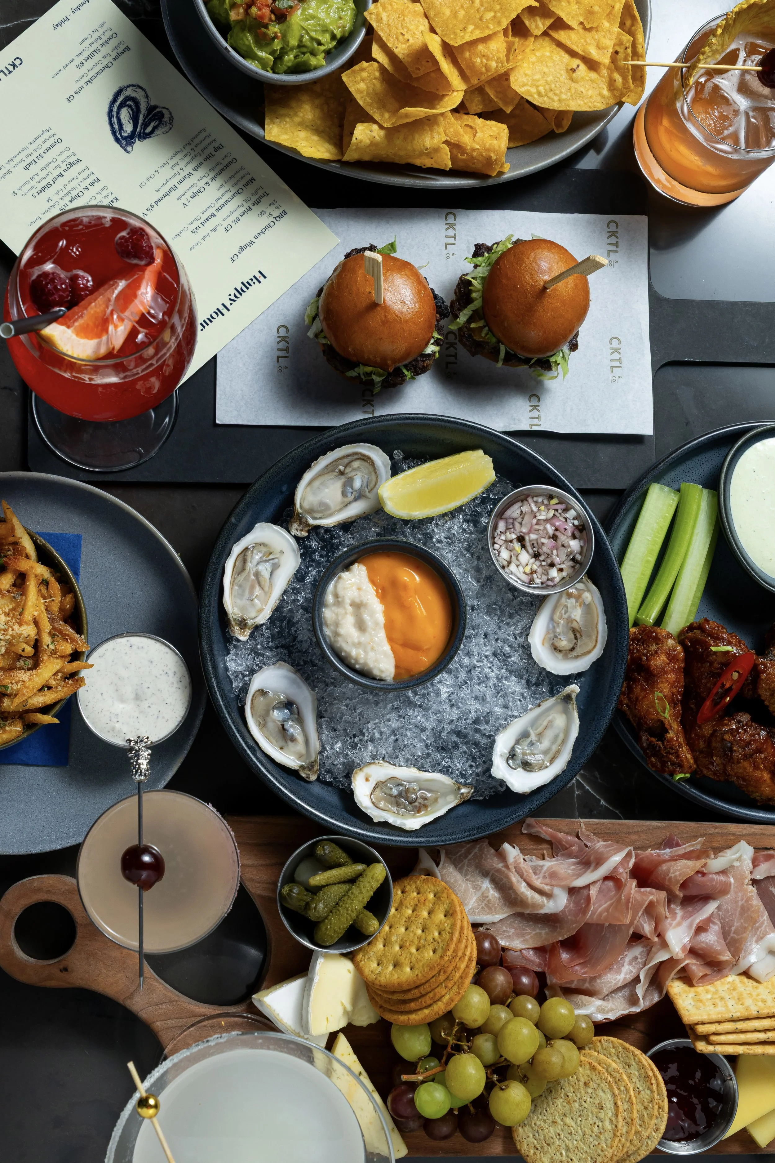 A table filled with various appetizers and drinks, including oysters on ice with lemon wedges, sliders with buns and lettuce, a fruit cocktail with raspberries and orange slices, cheese, grapes, crackers, cucumbers, and bowls of sauces and dips.