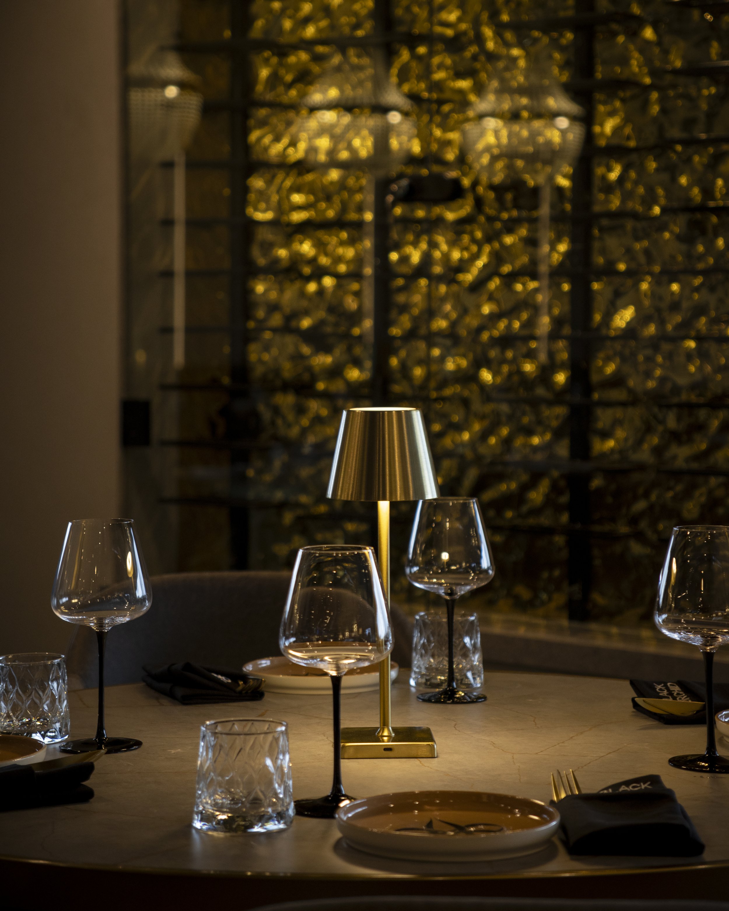 Elegant dining table setup with multiple wine glasses, a modern gold table lamp, and a dark background featuring a gold decorative wall. The table has a white marble surface with plates, napkins, and cutlery arranged.