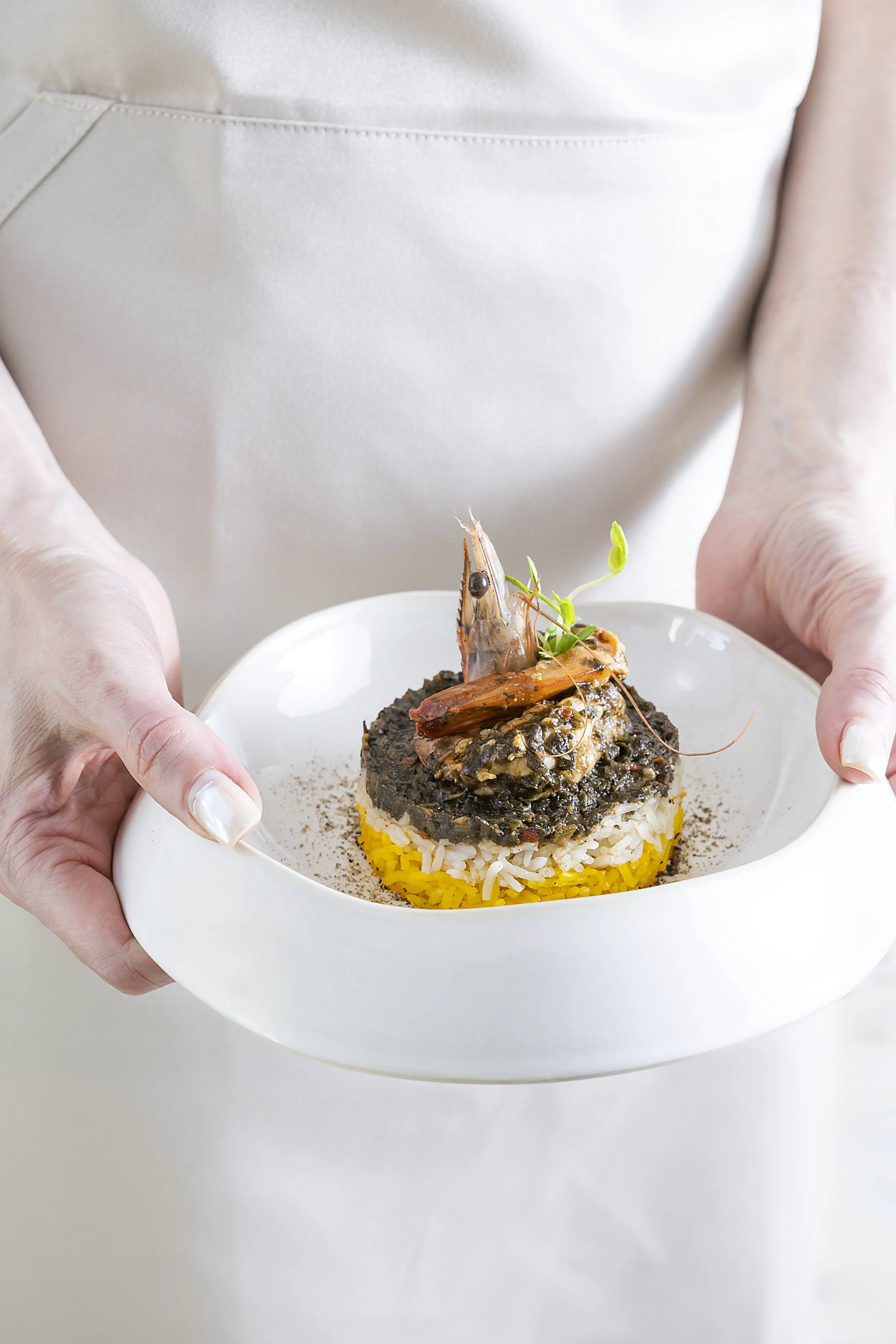 A person in a white apron holding a white plate with a layered dish topped with a shrimp and green garnish.