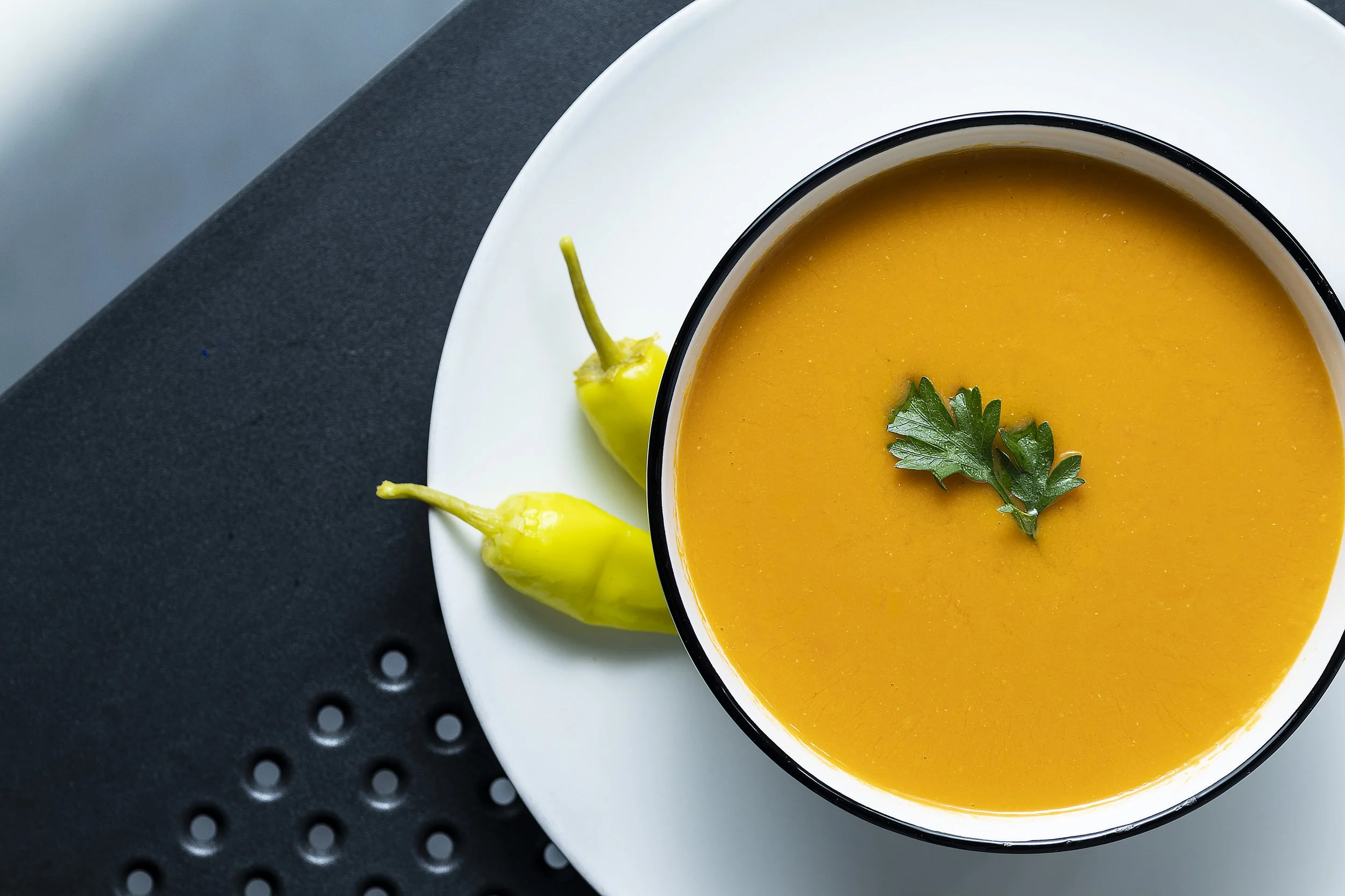 A bowl of orange soup garnished with green parsley, placed on a white plate with three yellow peppers nearby, on a black textured surface.