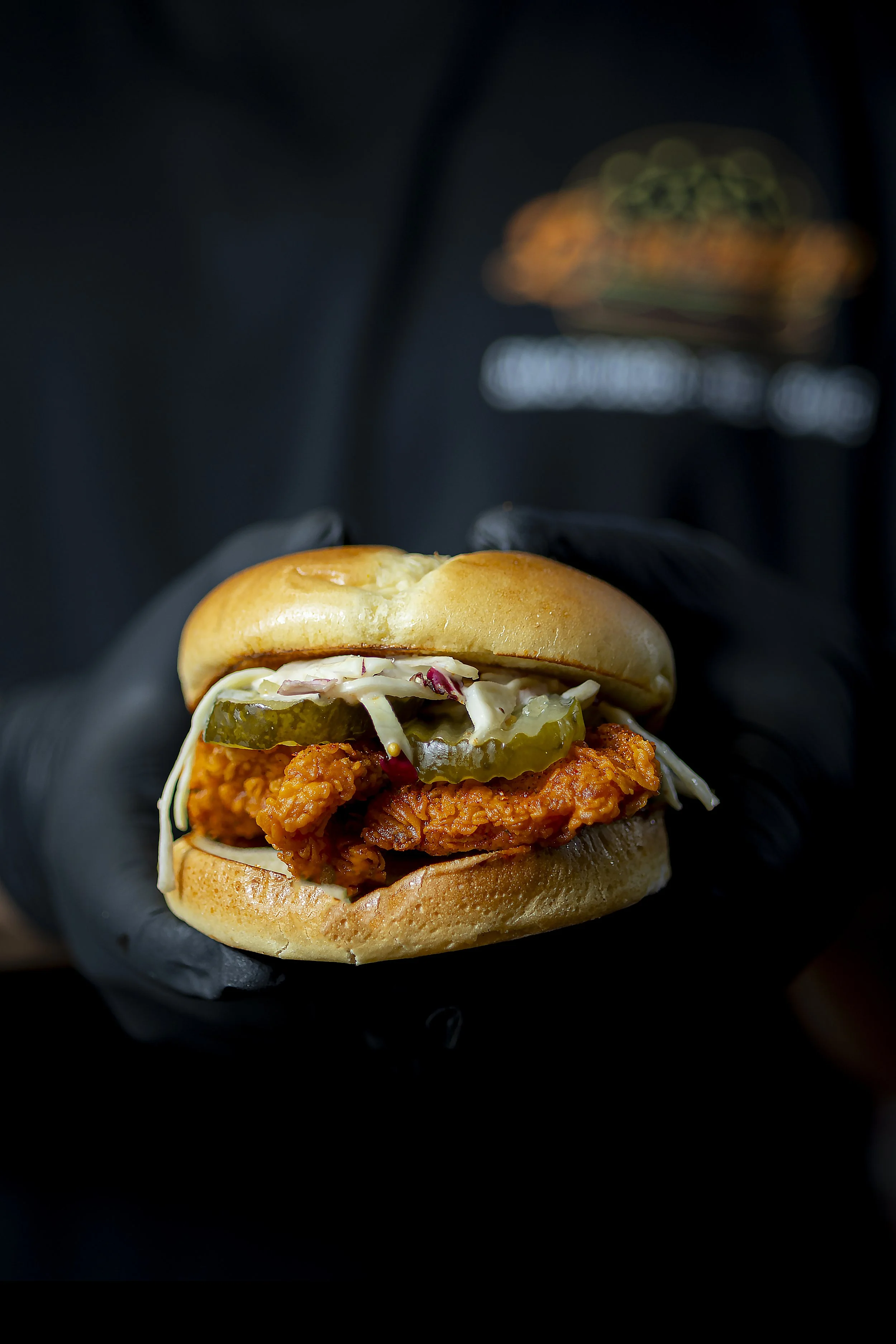 Close-up of a fried chicken sandwich in a bun topped with pickles, shredded lettuce, and coleslaw, held by a person wearing black gloves against a dark background with a blurred restaurant logo in the distance.