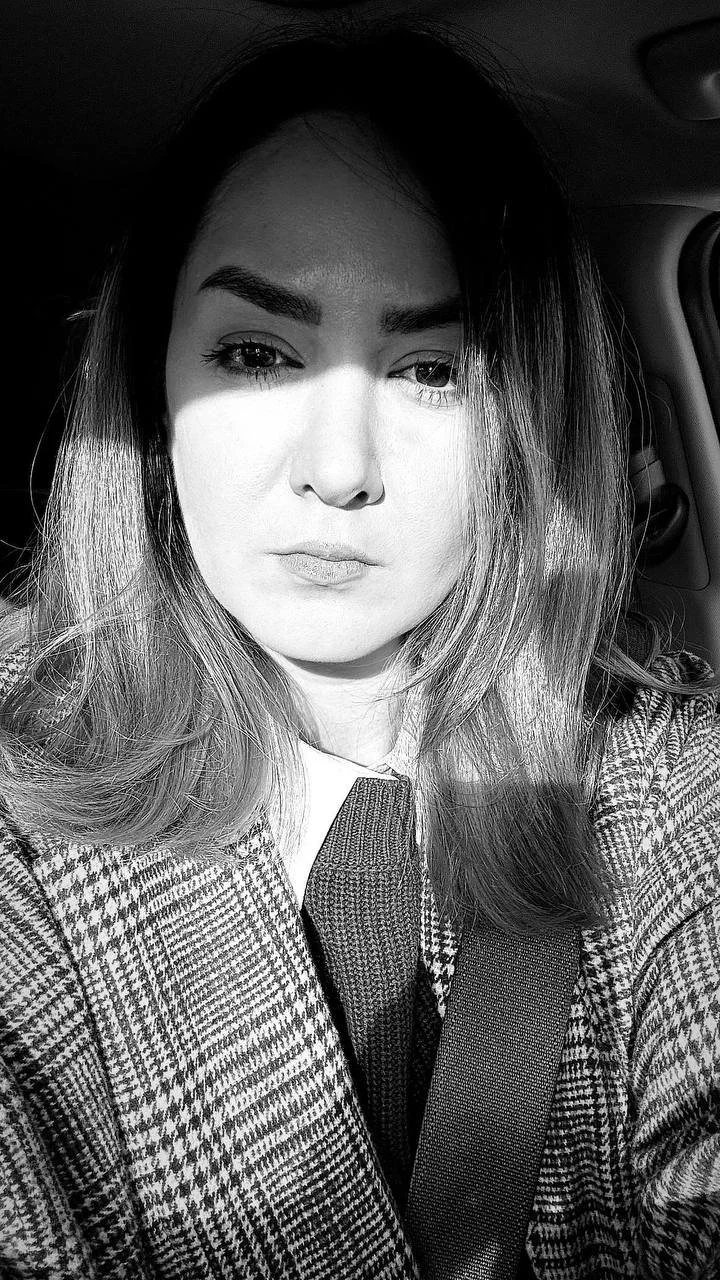A woman with straight hair and well-defined eyebrows sitting in a car, taking a selfie in black and white.