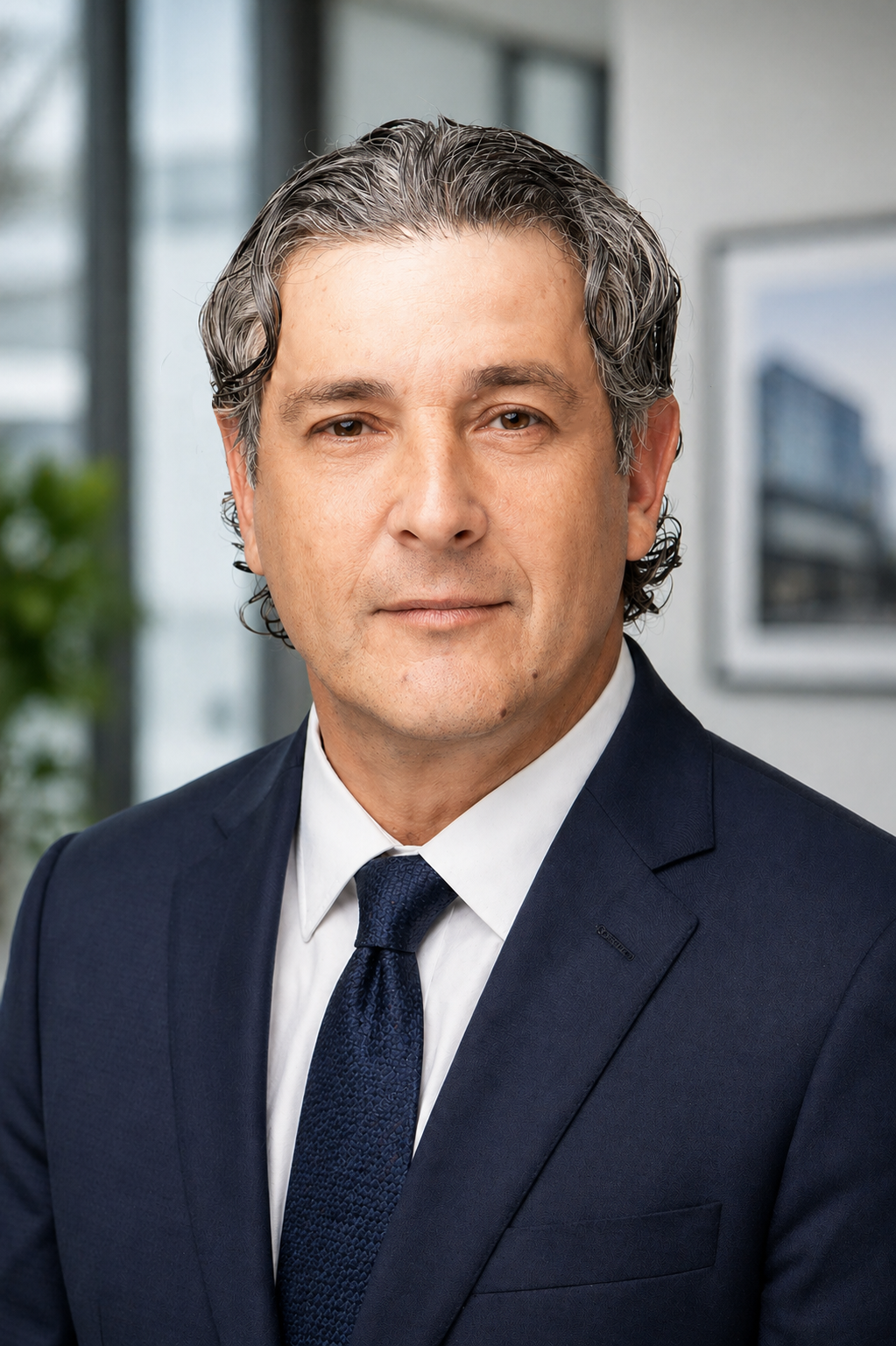 A professional man in a navy blue suit with a white shirt and dark blue tie, posing for a headshot in an office setting. The background shows large windows and framed artwork.