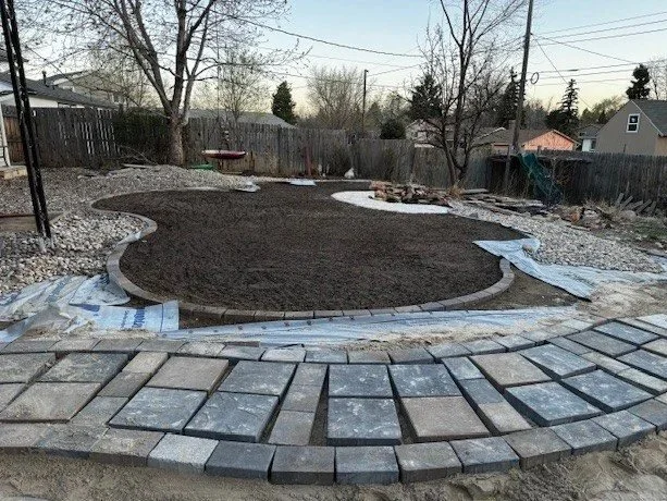 A backyard with a partially completed landscape project. It has a circular paved area with gray pavers in the foreground and a surrounding dark soil area, possibly for a lawn or garden.
