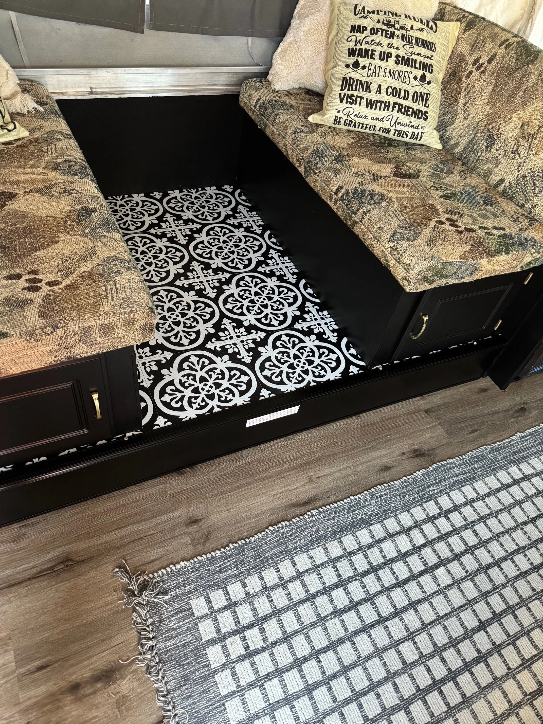 A cozy camper with a black and white patterned rug, two patterned upholstered couches, and a decorative pillow with nature-inspired text. The area features wood flooring and a window with curtains.