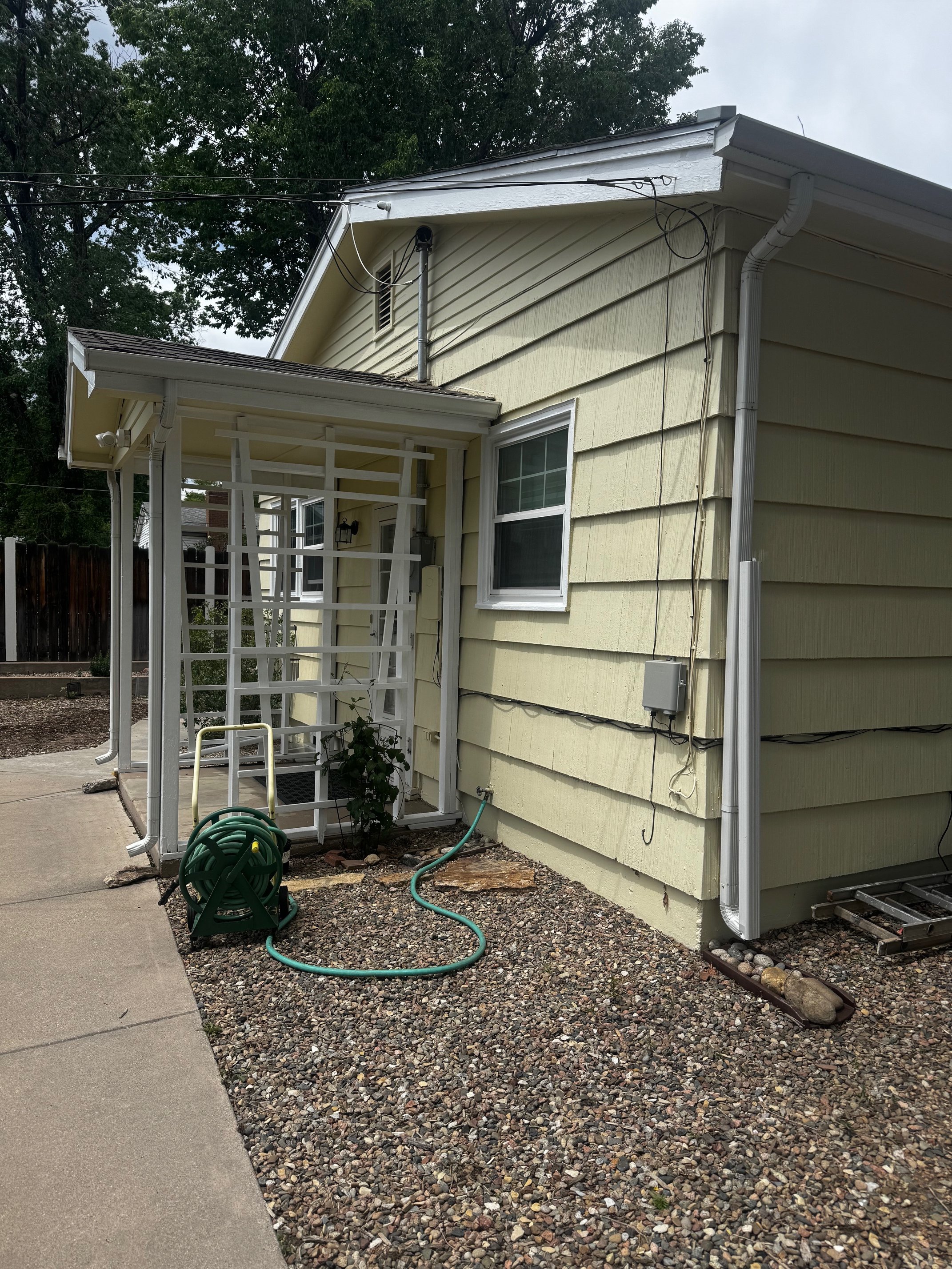 The side view of a yellow house with white trim and a small porch area, featuring a tan sliding window, some electrical wiring, and a garden hose on the gravel ground.