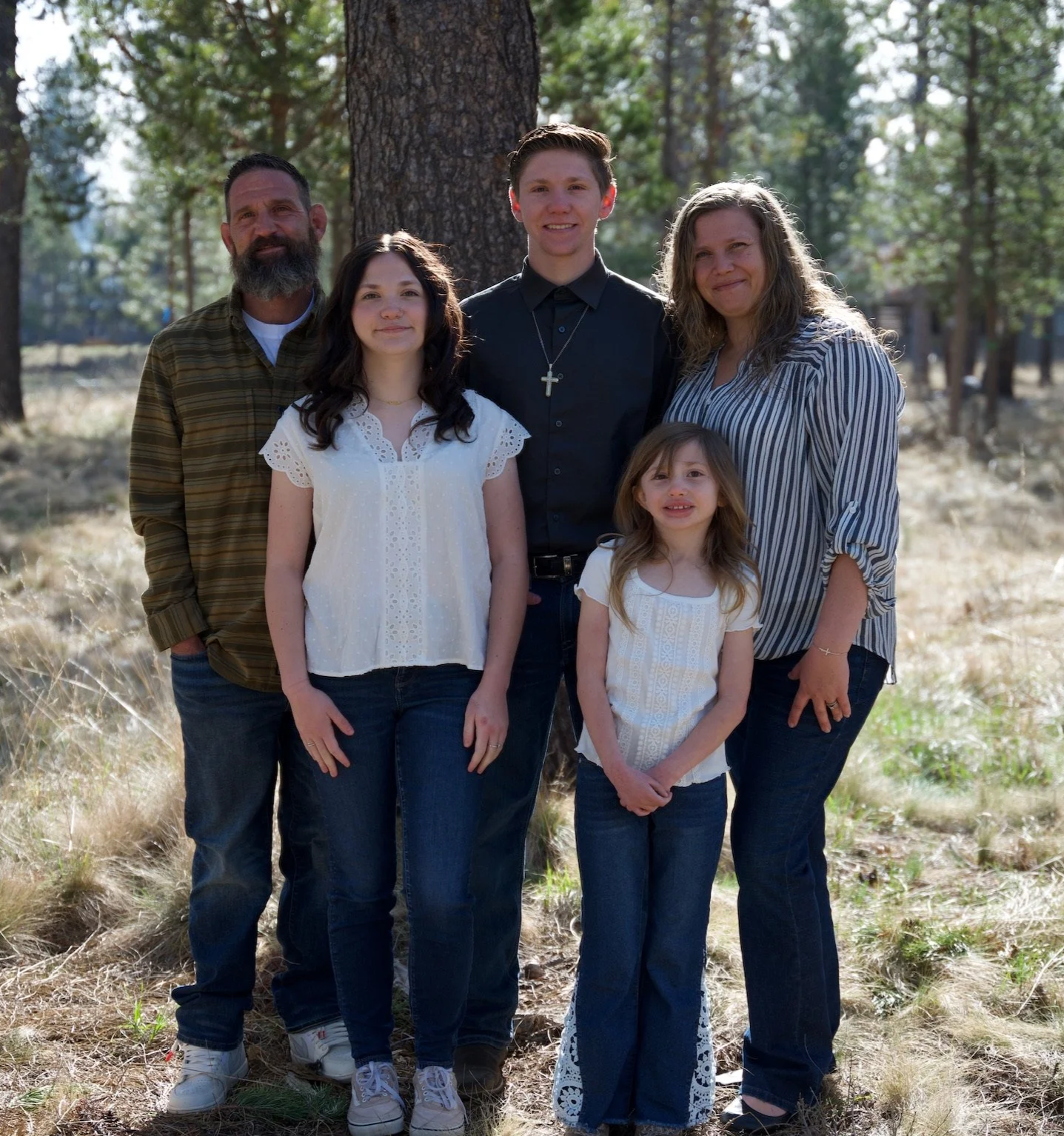 Family photo of five people standing outdoors in a wooded area, smiling at the camera.
