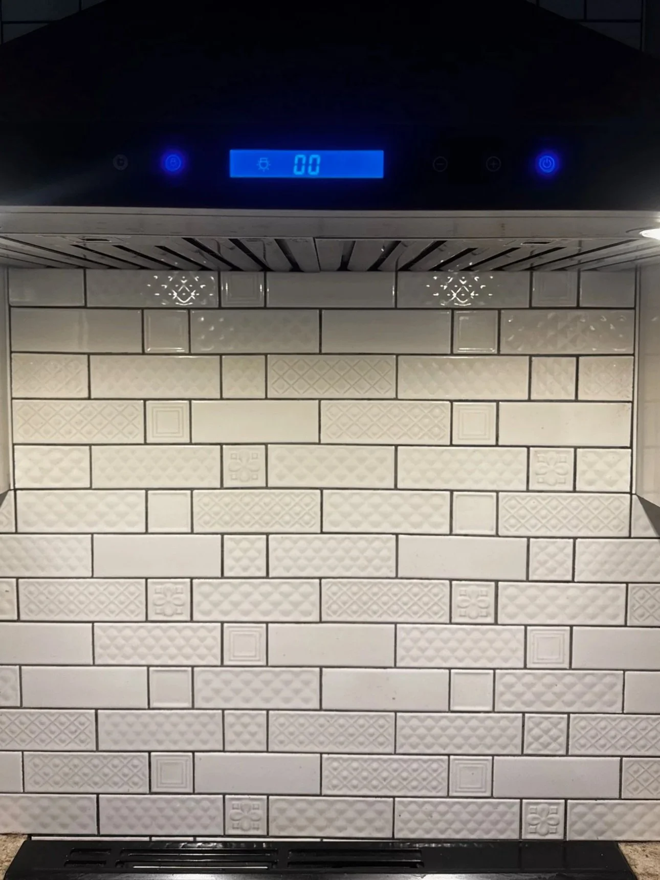 A kitchen range hood with digital display and control buttons above a white textured tile backsplash.