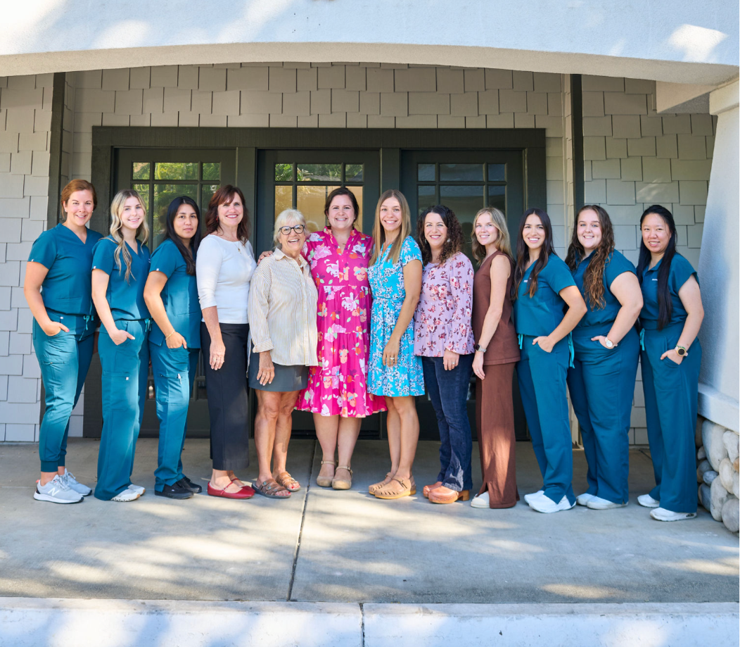 A group of twelve women standing in front the clinic, some in medical scrubs and others in casual and business attire, smiling for a photo.
