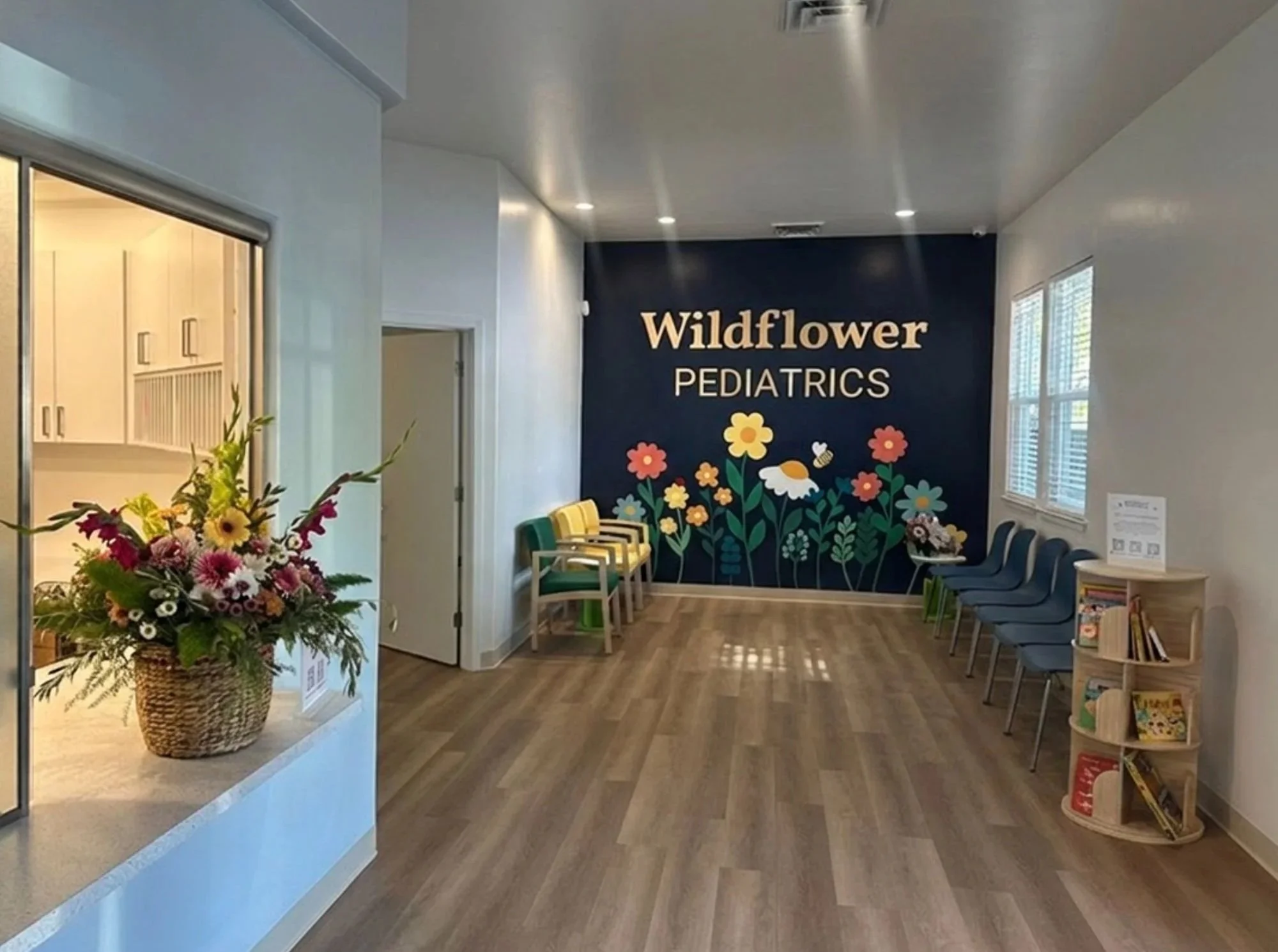 Interior of pediatric clinic with a dark blue accent wall decorated with colorful flowers, a sign reading 'Wildflower Pediatrics,' and a row of chairs for waiting patients. There are windows with blinds, a floral arrangement in a wicker basket, and a small bookshelf with children's books.