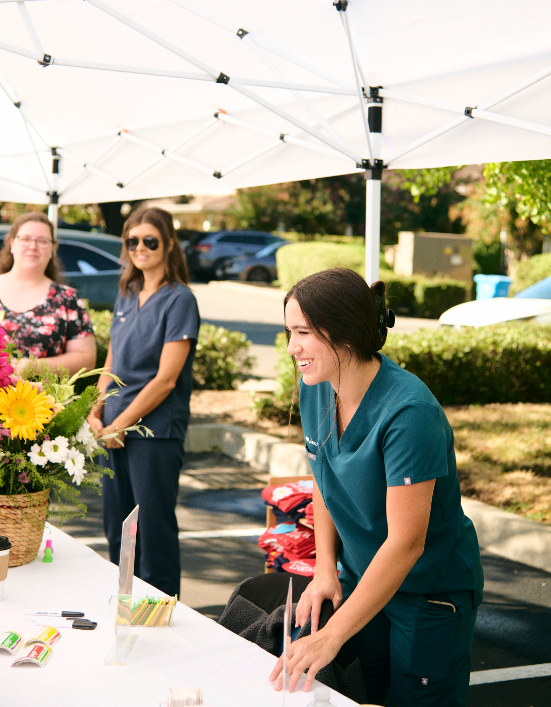 A smiling woman in teal scrubs arranging items on a table outdoors under a white canopy, with two women in the background, one in blue scrubs and the other in a floral shirt, standing behind a flower arrangement.