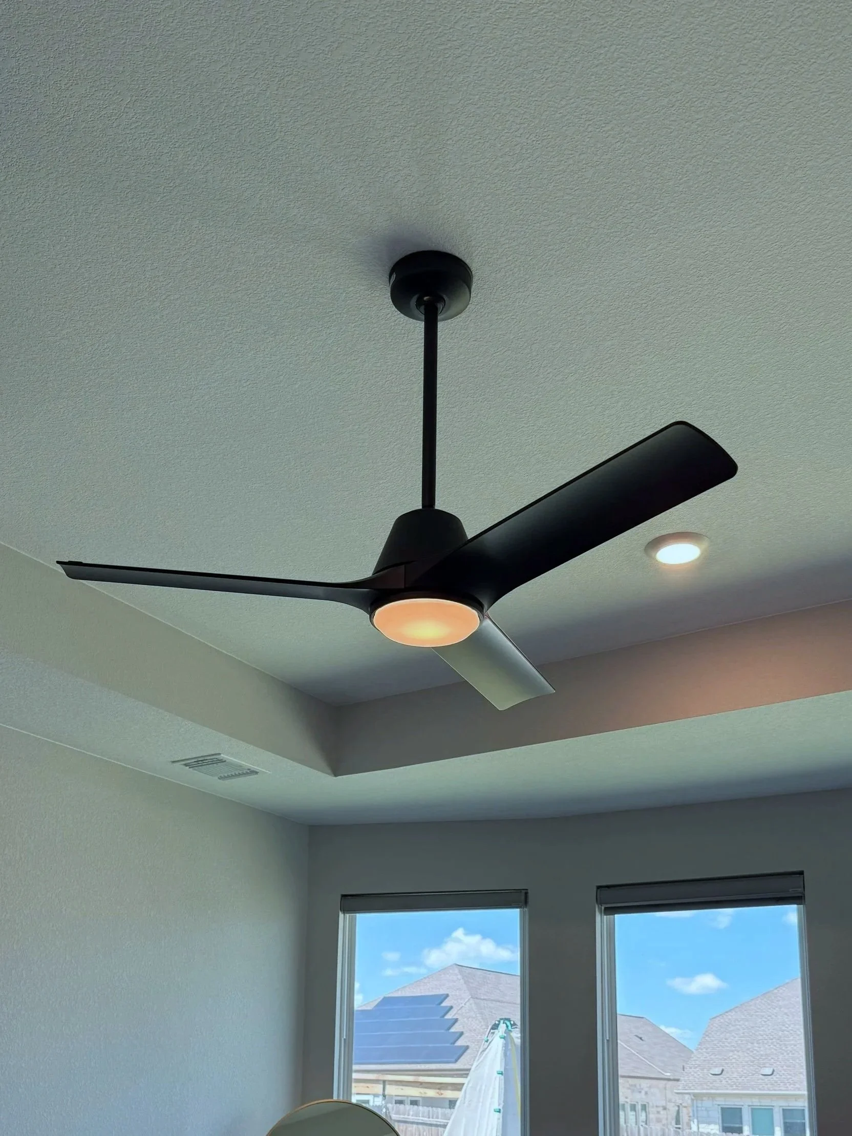 Ceiling fan with black blades and a light fixture mounted on a white textured ceiling, next to a window in a room with light-colored walls.