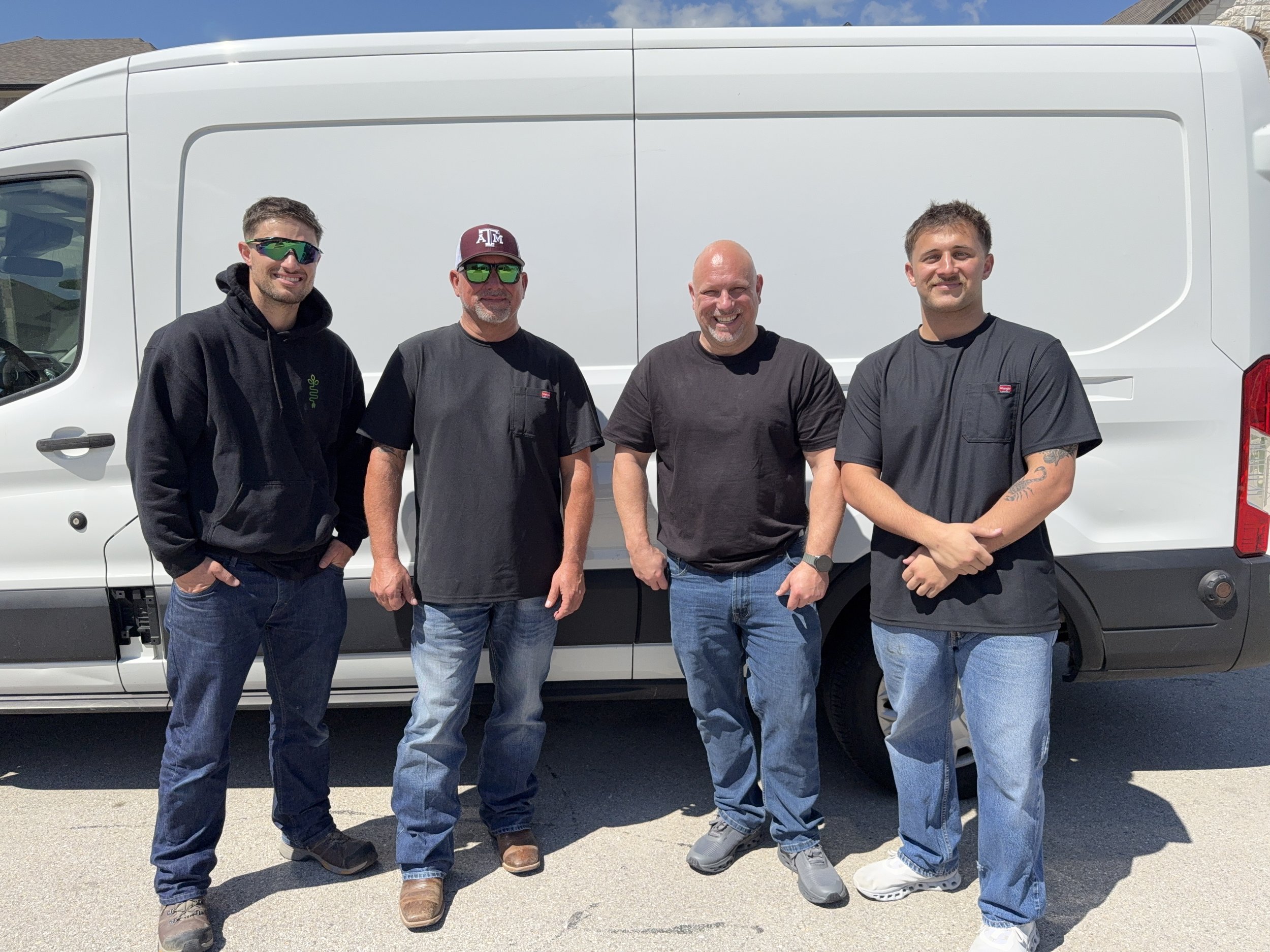 Four men standing in front of a white van, smiling at the camera outdoors on a sunny day.