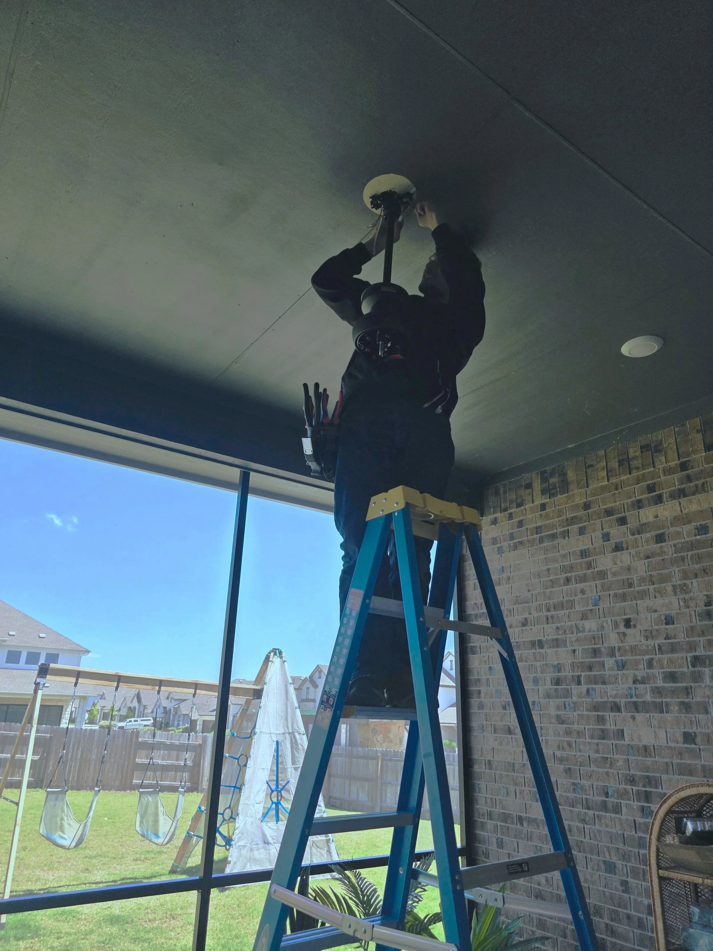 A person standing on a blue ladder, installing or repairing a ceiling light fixture in a room with large windows and a brick wall, with a backyard and houses visible outside.