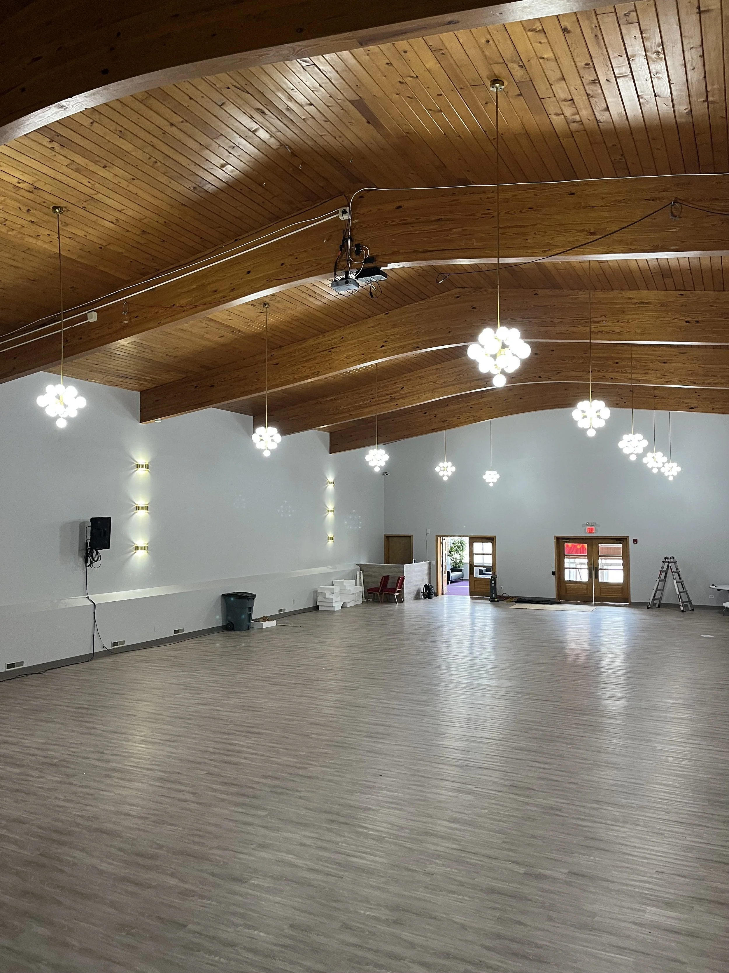 A large, empty room with wood-paneled arched ceiling, modern chandelier lights, and light-colored vinyl flooring. There are some chairs, boxes, and equipment near the back wall, which has double doors and a small window.