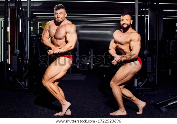 Two shirtless men with muscular physiques posing in a gym.