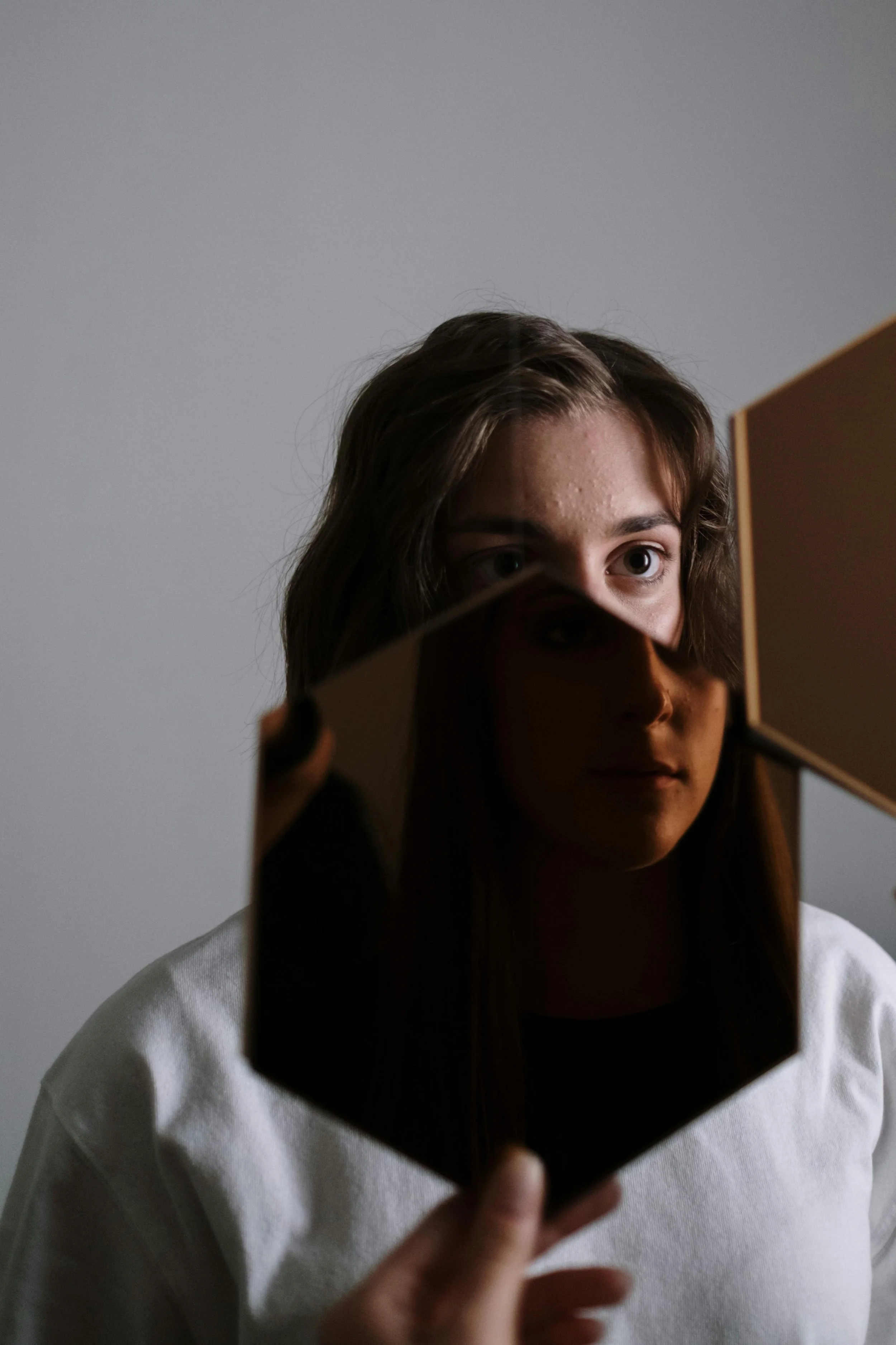 Young adult with long brown hair and white shirt holds small mirror in front of her face, reflecting only their eyes and forehead against plain, light gray background. She is contemplating her identity.