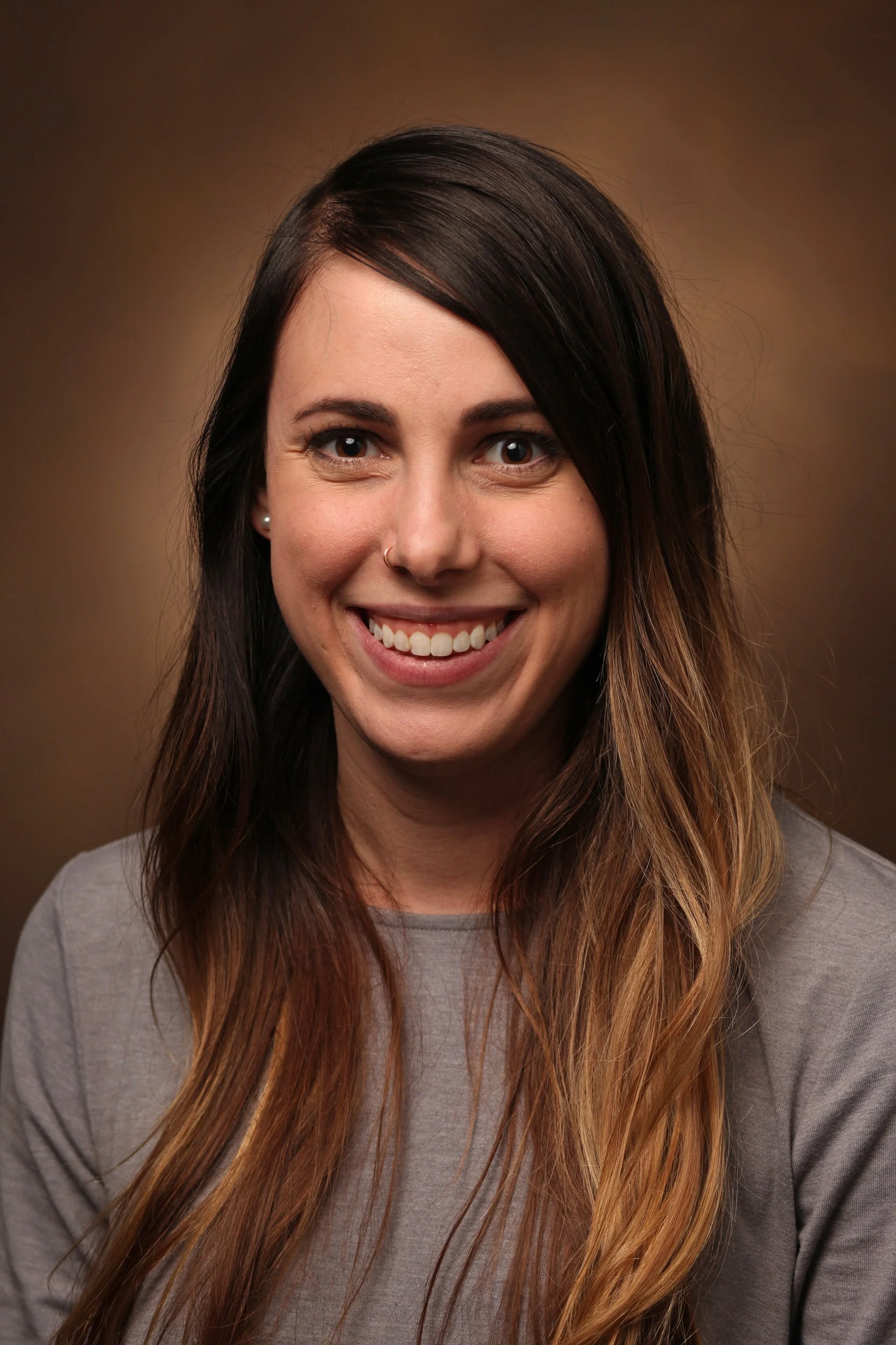 A woman with long, wavy hair, wearing a light gray top, smiling, with dark brown hair and a brown gradient background.