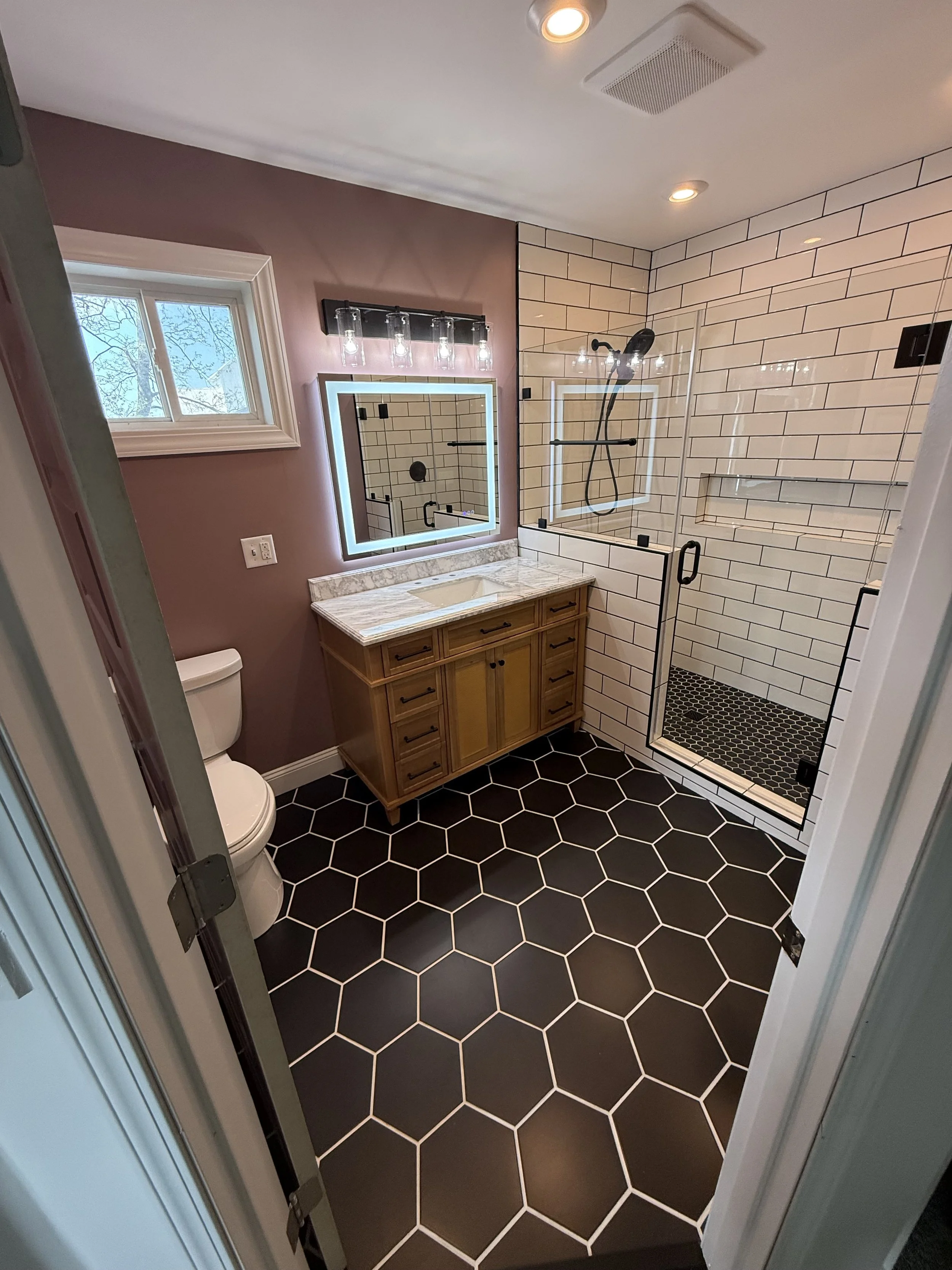 Bathroom with black hexagon floor tiles, a toilet, wooden vanity with marble countertop, illuminated mirror, window with tree view, and a walk-in shower with white subway tiles and black fixtures.