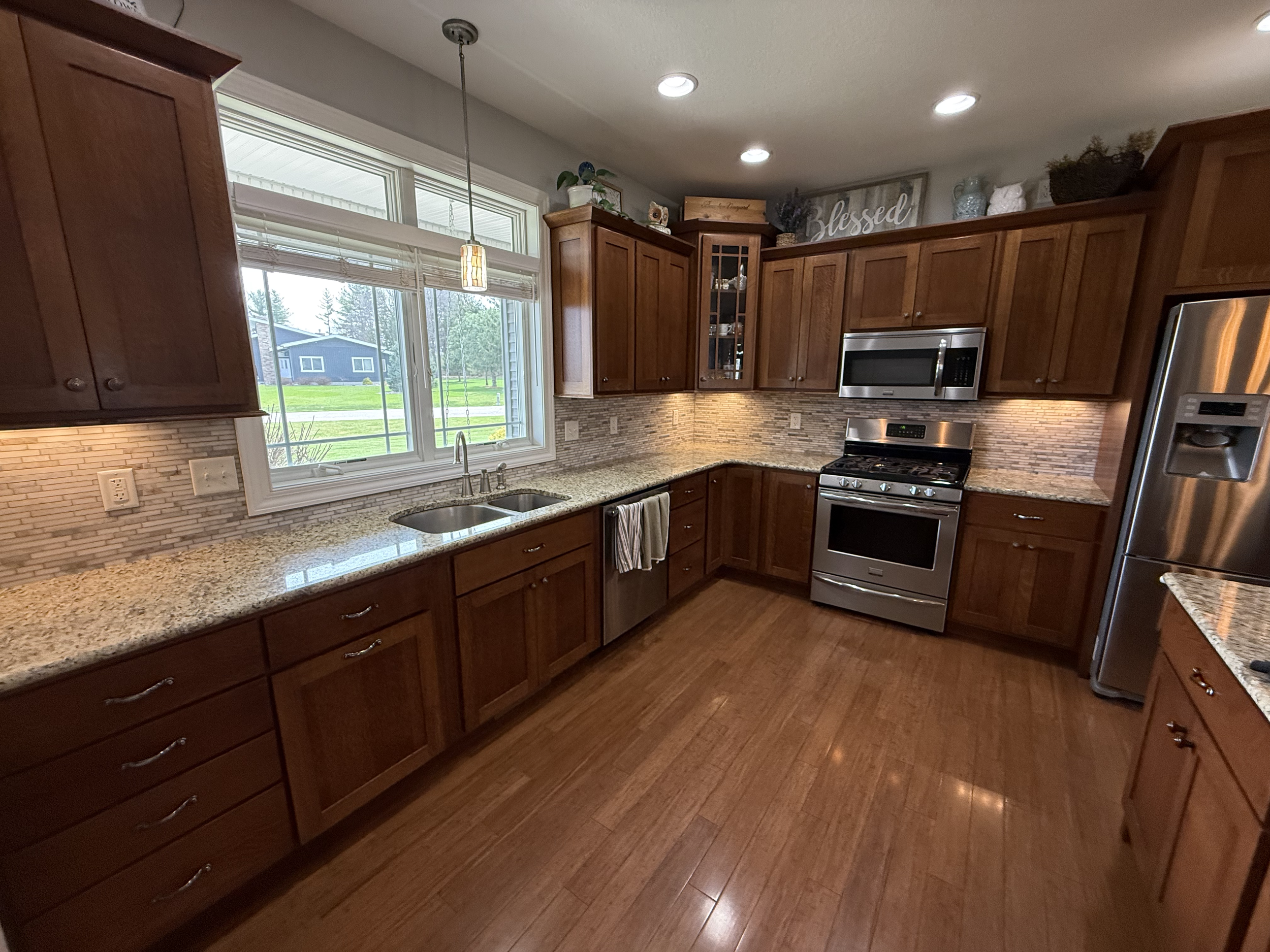 Kitchen with wooden cabinets, granite countertops, stainless steel oven, microwave, and refrigerator, large window with blinds, and decorative items on top of cabinets.