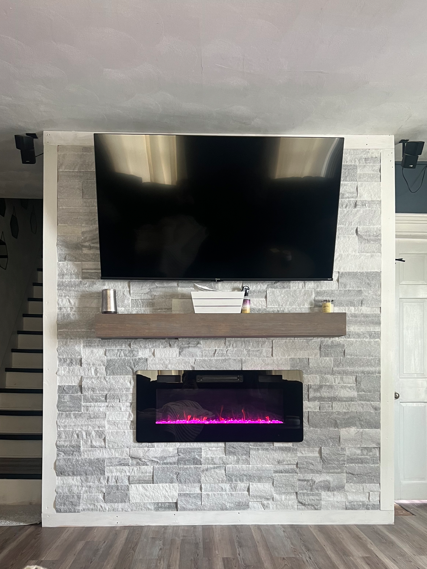 Living room feature wall with mounted flat-screen TV above a modern electric fireplace with pink flames, flanked by a stone veneer wall and white trim, with a wooden mantel and small decorative items below.