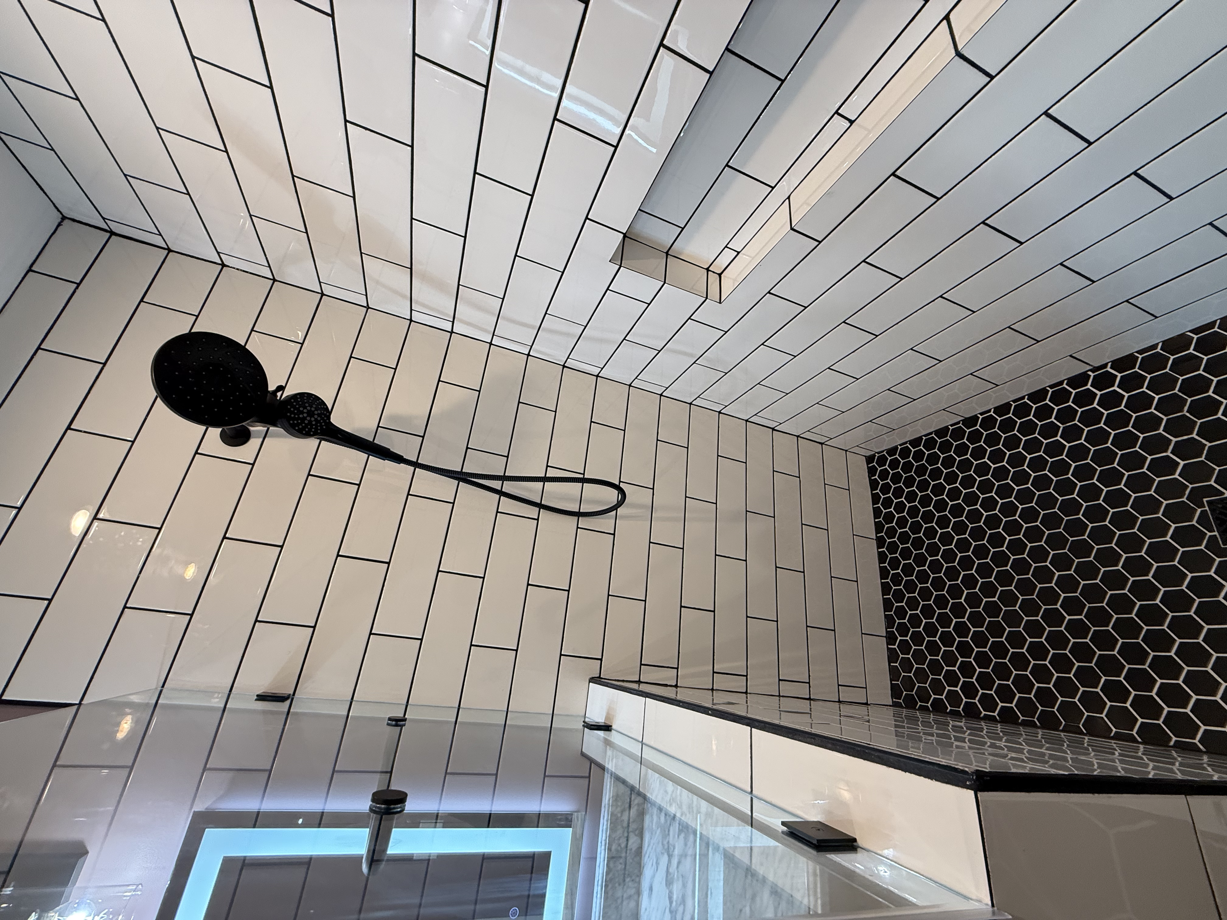 This image shows a bathroom with white rectangular subway tiles on the wall, a black showerhead attached to a flexible hose, and a black hexagonal tiled floor. There is a strip light on the ceiling and a glass door leading into the bathroom.