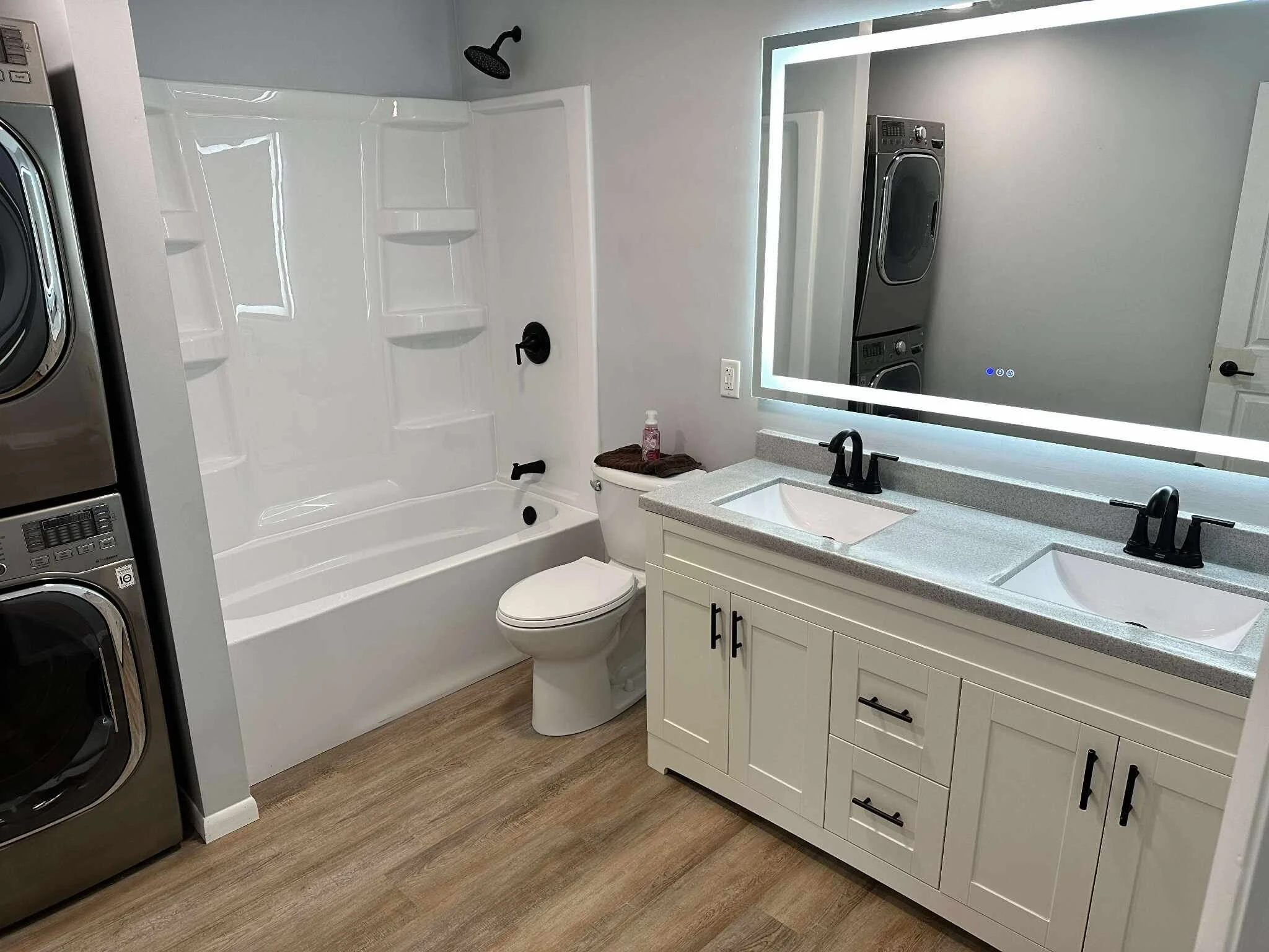 Modern bathroom with a bathtub and shower, a toilet, a white vanity with double sinks, a large illuminated mirror, and laundry appliances visible in the reflection.