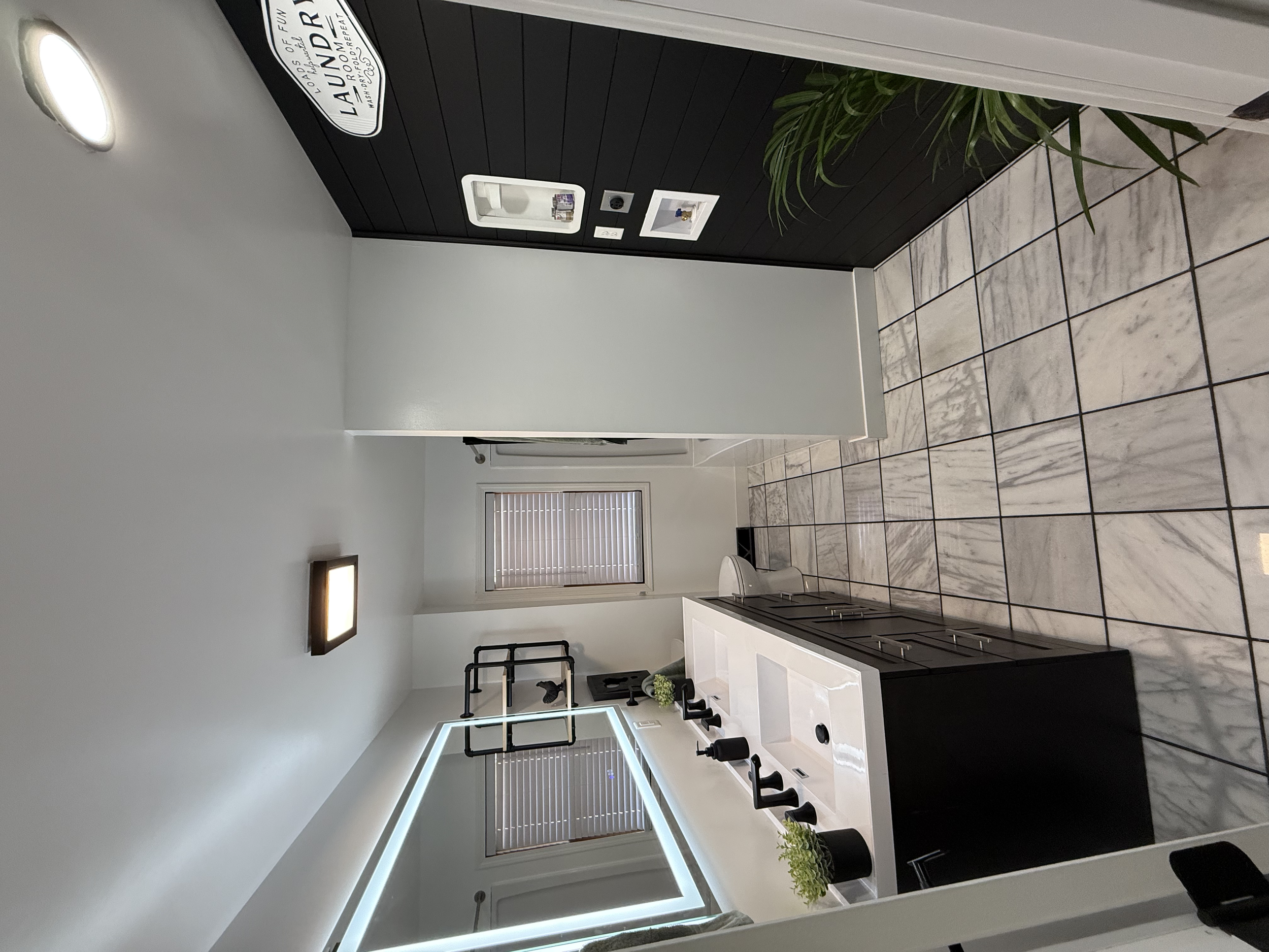A modern bathroom with black and white marble tile flooring, dark wall paneling, a white vanity with a black cabinet, a mirror with LED lighting, a window with blinds, and a towel holder.