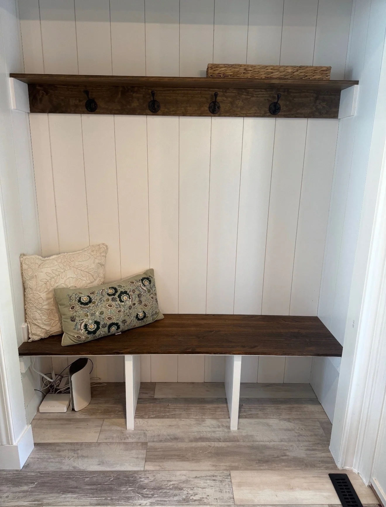 Indoor entryway with a wooden shelf and hooks on a white wall, two decorative pillows on a wooden bench, and a floor vent.