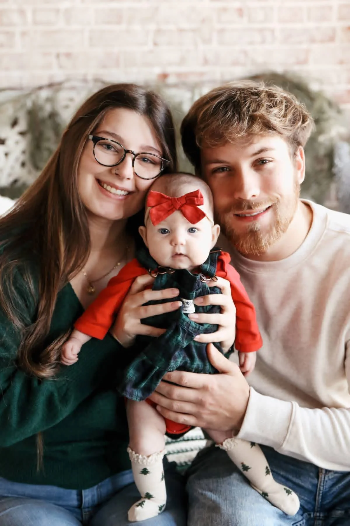 A smiling woman with long brown hair, glasses, and a man with short blond hair and a beard holding a baby with a red bow on her head, dressed in festive red and dark green clothing, in front of a brick wall.