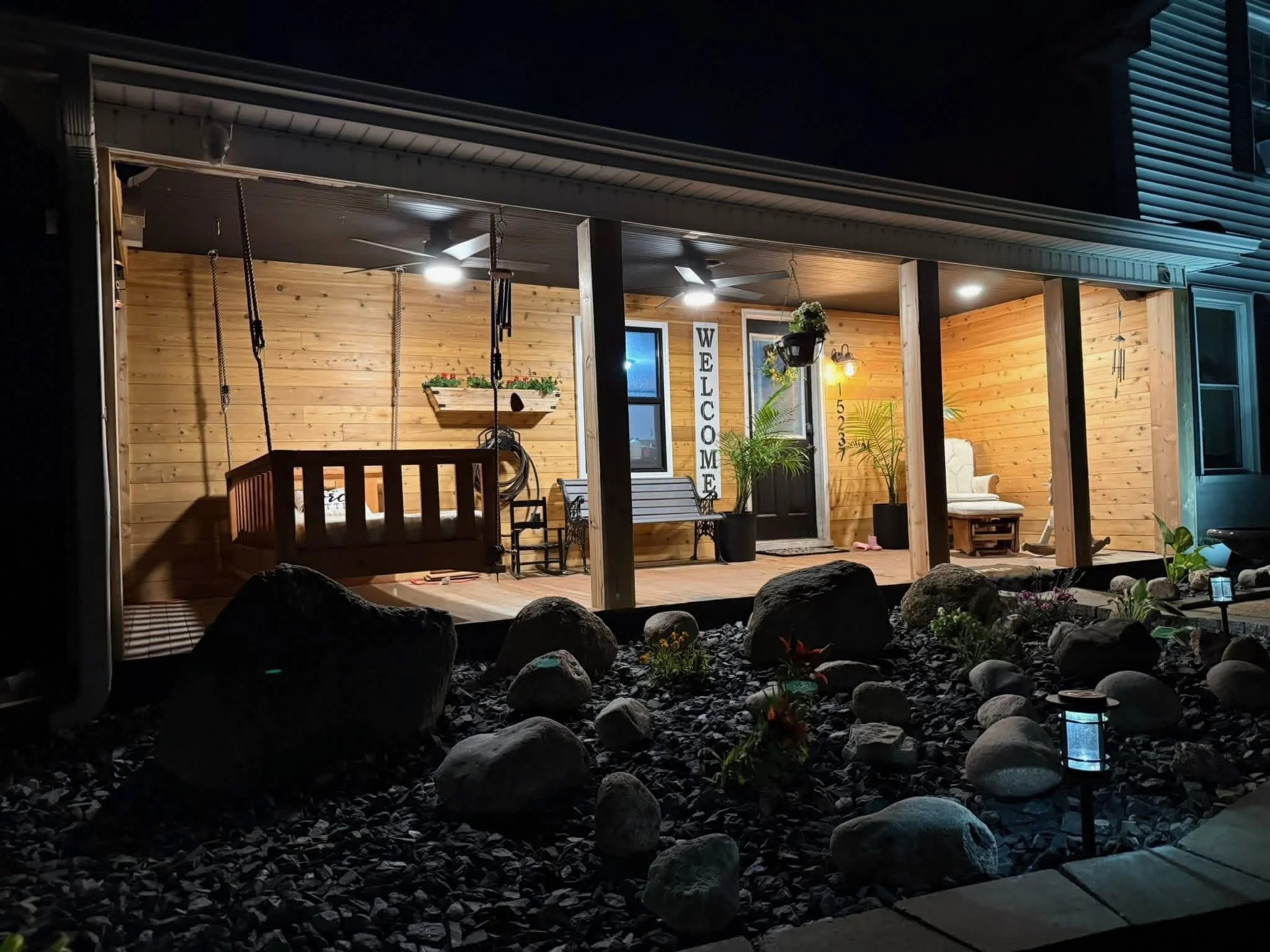 Night view of a covered porch with wooden walls, ceiling fans, hanging plants, sitting area, and a welcome sign, illuminated by outdoor lights, and surrounded by rocks and plants.