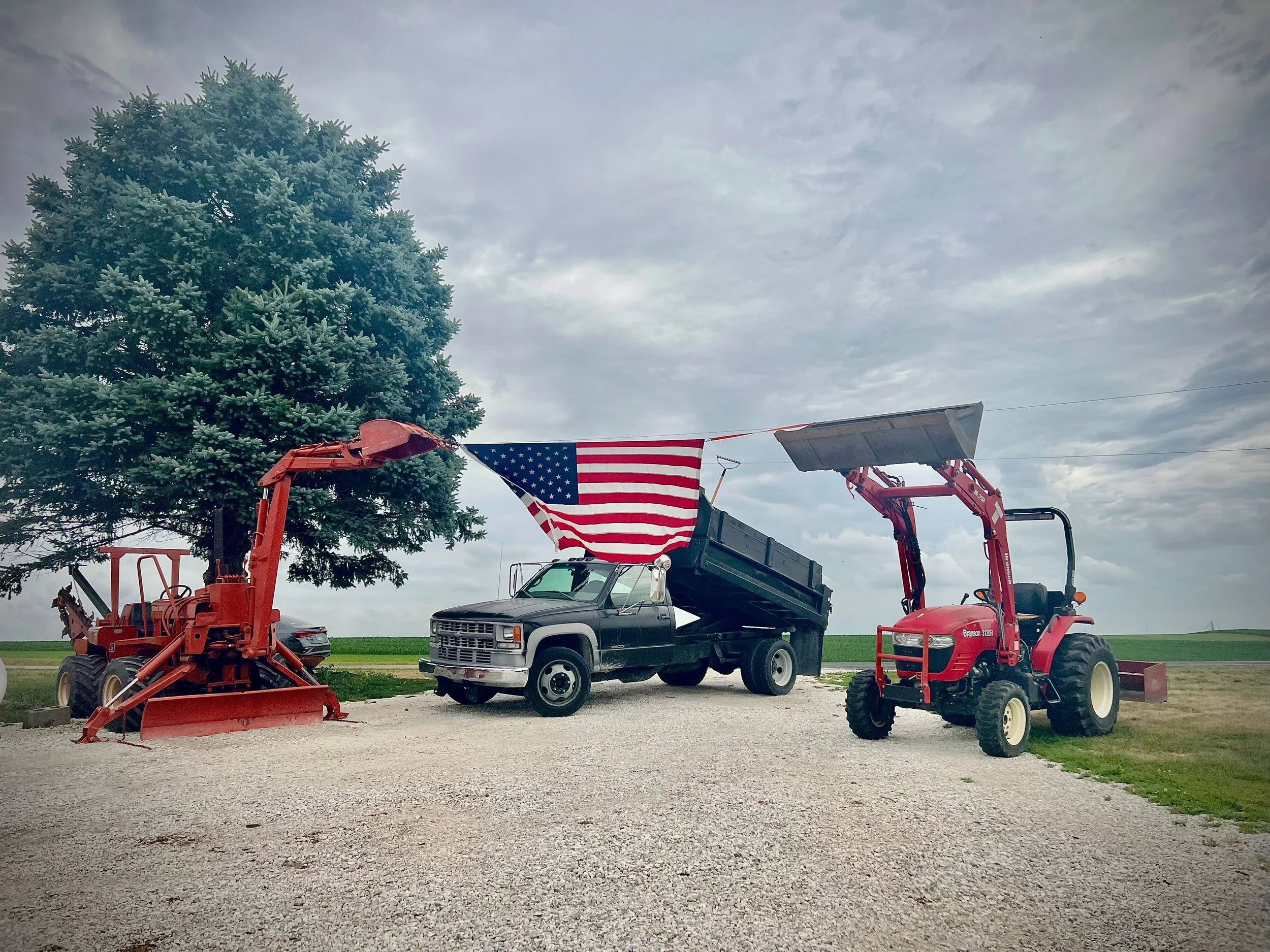 A large evergreen tree next to antique farming equipment, a black pickup truck, and an American flag on a pole under a cloudy sky.