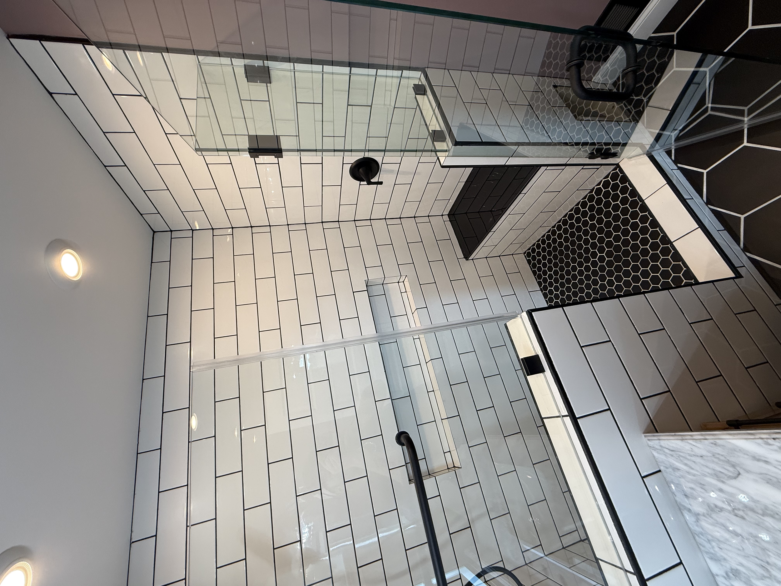 Interior view of a modern building with tiled walls in white and black, black handrails, and a glass door with black handles, floor with black hexagon tiles, and ceiling lights.