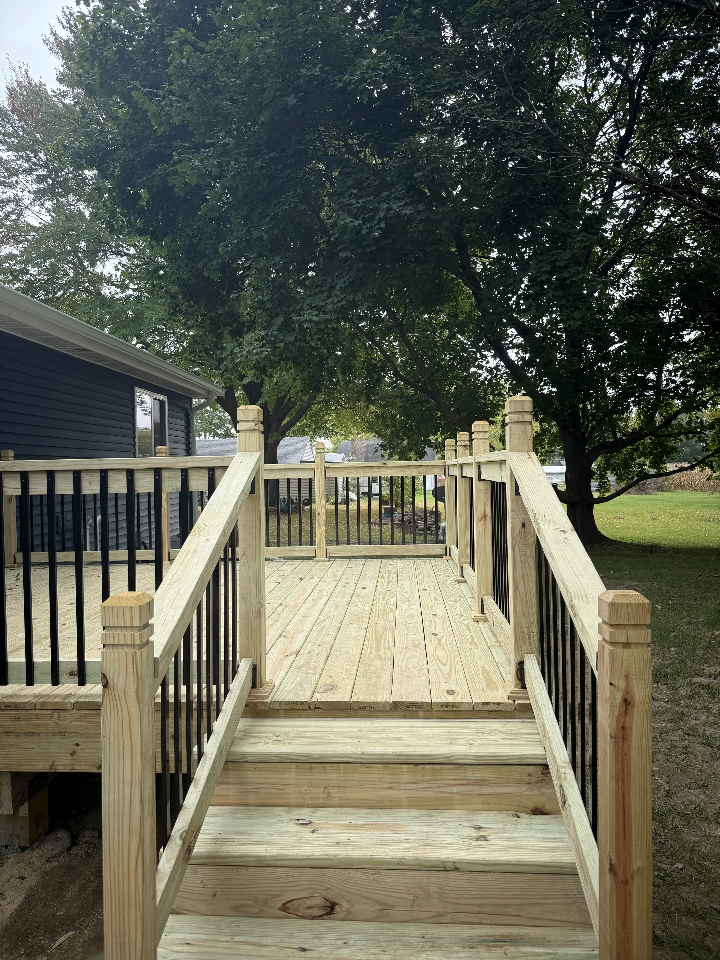 A newly constructed wooden deck with stairs and black metal railing in a backyard with large trees and a blue house.