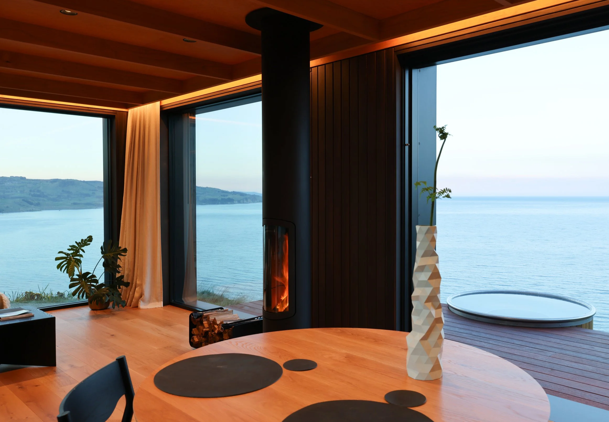 Living room with large floor-to-ceiling windows showing a scenic view of water and hills. Wooden ceiling and walls, a round wooden table with black placemats, a white geometric sculptural tall vase, plants, and a suspended fireplace.