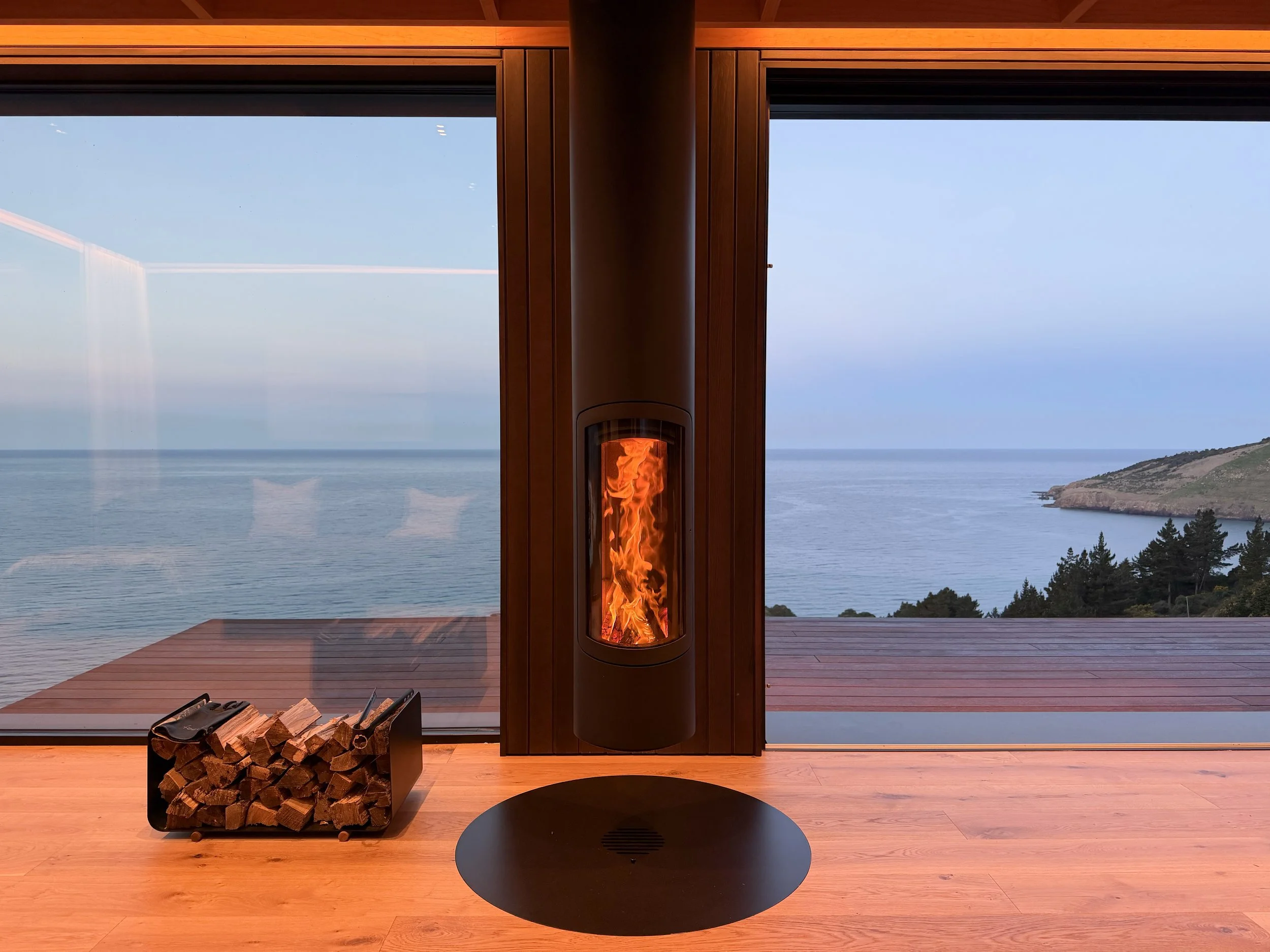 A suspended fireplace with a fire burning, located in front of large windows overlooking an ocean view, wooden floor, firewood stacked nearby.