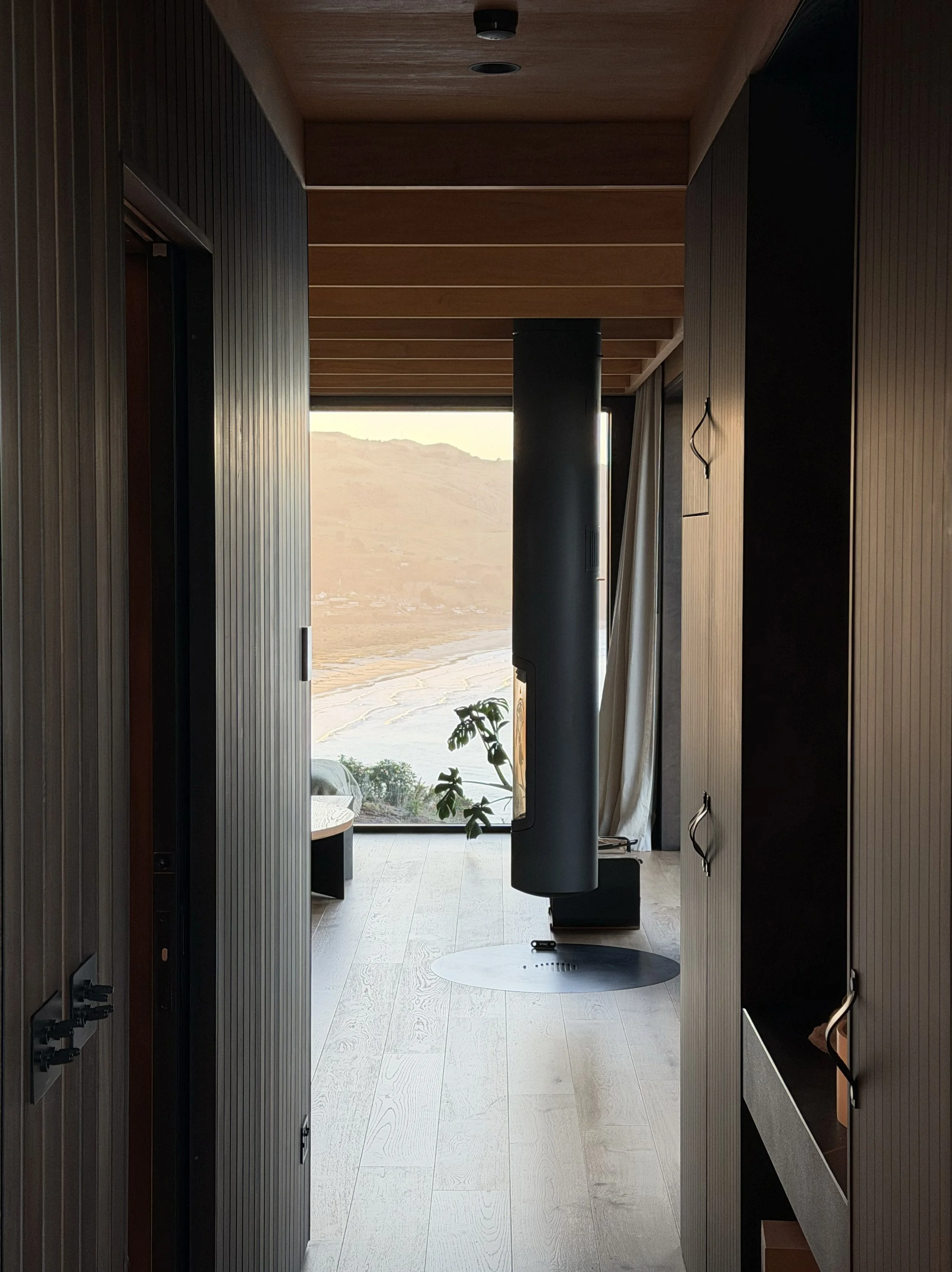 Interior view of a modern clifftop cabin with wooden walls and ceiling, large glass windows, a black suspended fireplace.