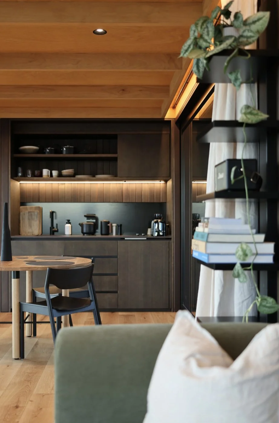 A modern kitchen with dark cabinetry, open shelves with dishes, a wooden ceiling, and a cozy dining area with a wooden table and black chairs. A green plant is on a black shelf beside a white curtain in front of a black window.