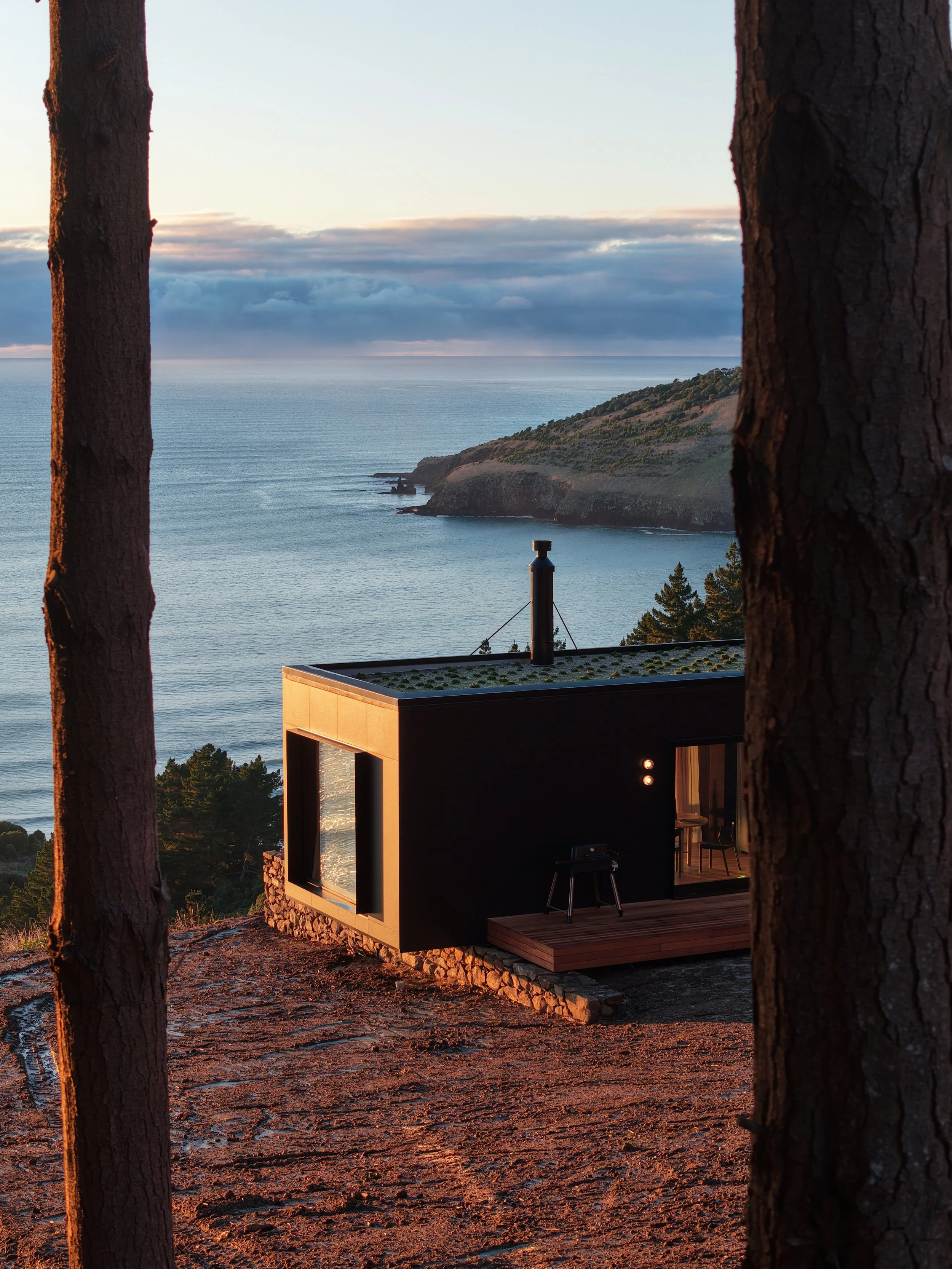 Black cabin with large windows, overhanging a rock wall, with views over the ocean and cliffs in the distance. The house is partially obscured by two large trees in the foreground, with a sunset sky and clouds above.