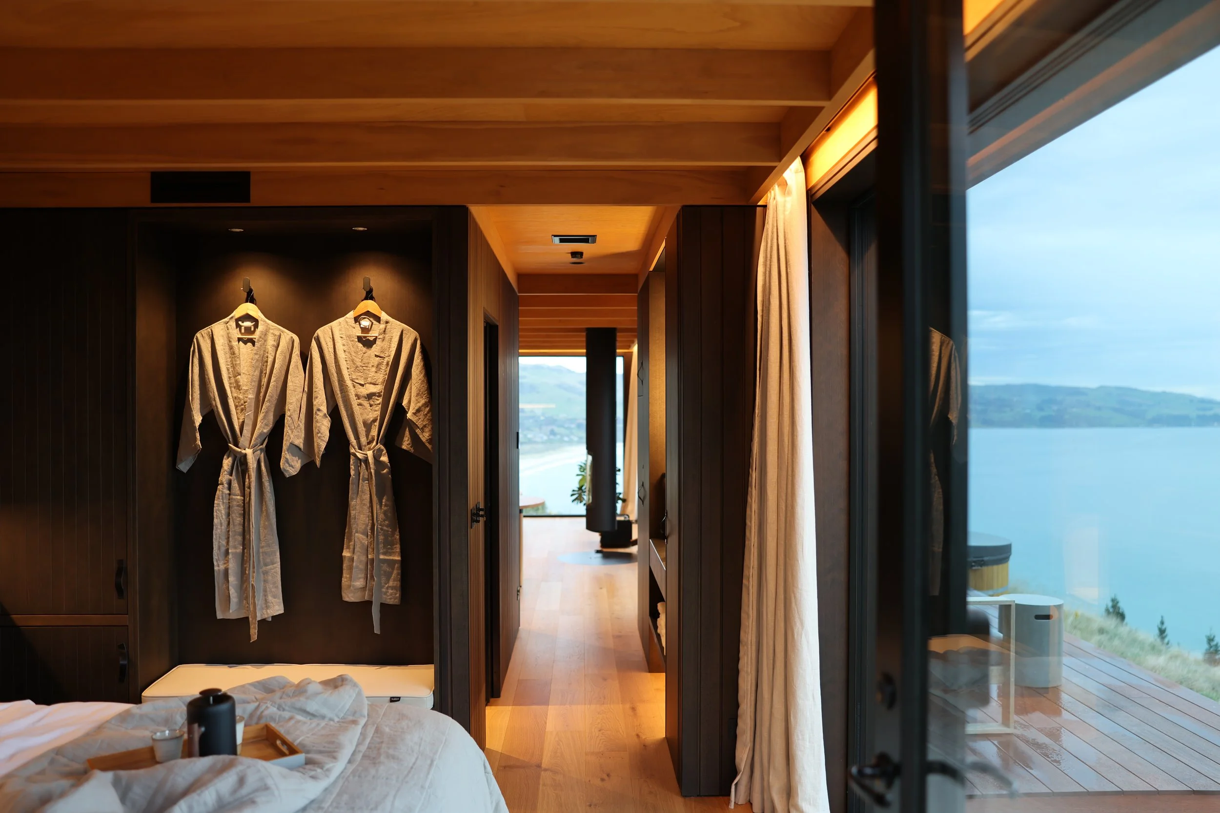 Bedroom with white unmade bed and black wardrobe with hanging robes, wooden ceiling and floor, large window with outdoor view of ocean and hills.