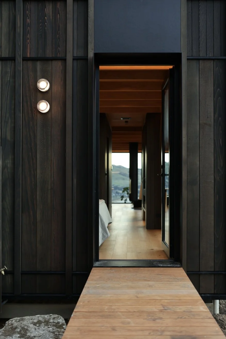 View of a doorway leading into a black cabin with dark wooden exterior walls and a wooden walkway, revealing a room with large windows and scenic ocean views.