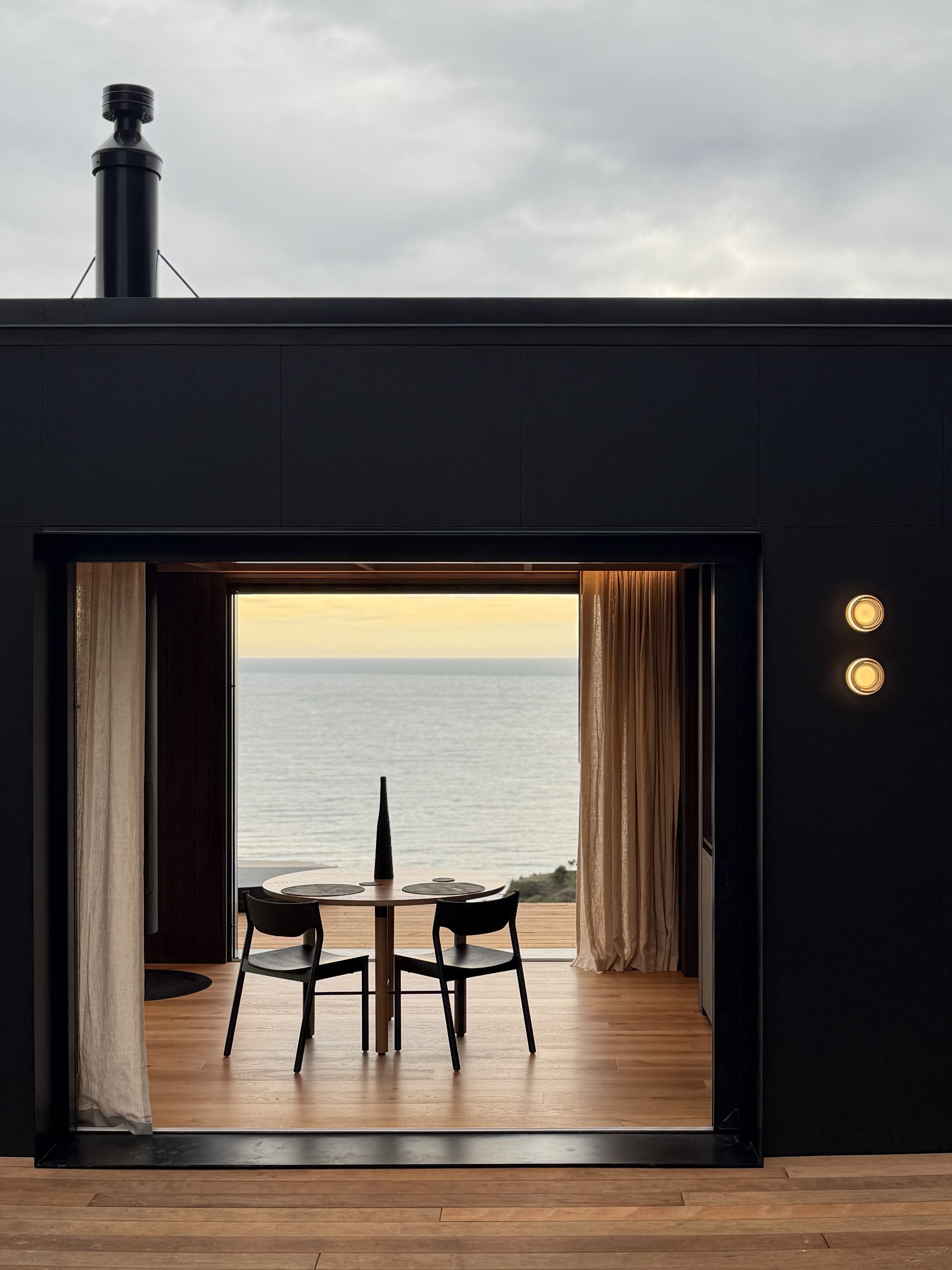 View through a clifftop cabin, with a dining table overlooking the ocean under a cloudy sky.