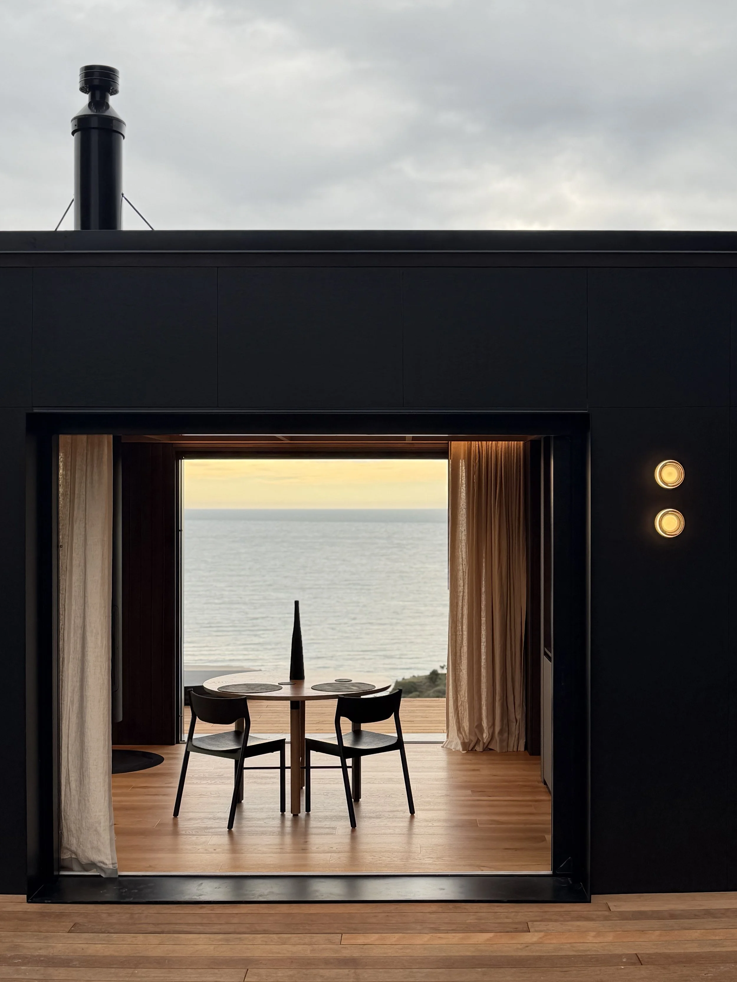 View through a clifftop cabin, with a dining table overlooking the ocean under a cloudy sky.