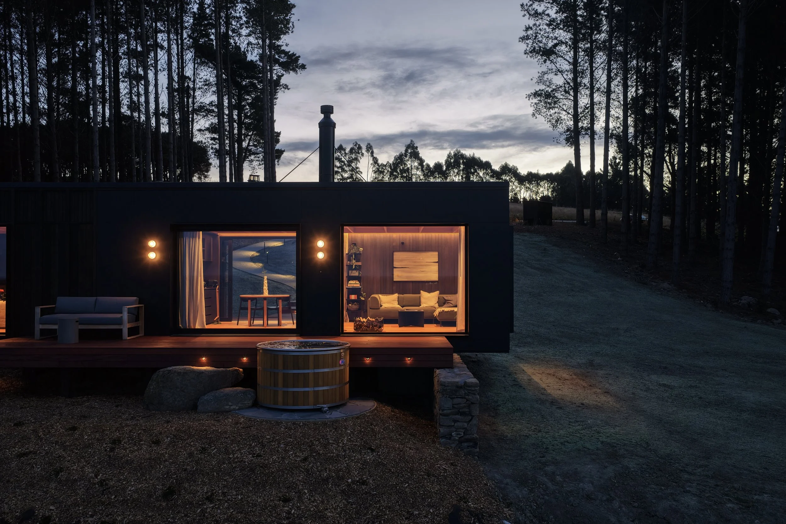 A modern cabin with large glass sliding doors illuminated from inside, and an outdoor hot tub, surrounded by a forest at dusk.
