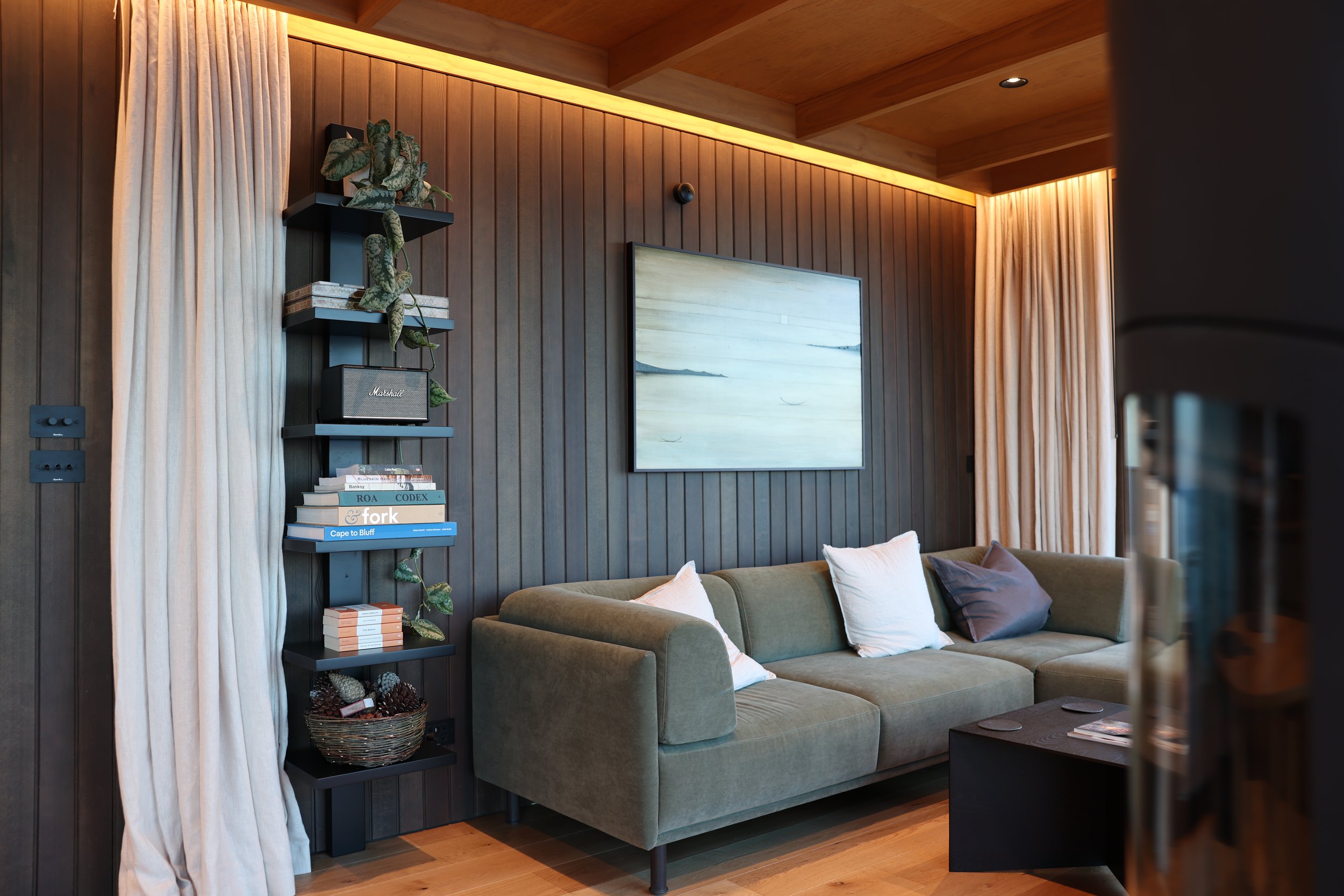 Living room with wooden paneled wall, beige curtains, a wall-mounted framed artwork depicting a coastal scene, a bookshelf with books and decorative items, and a green upholstered sofa with multiple pillows.