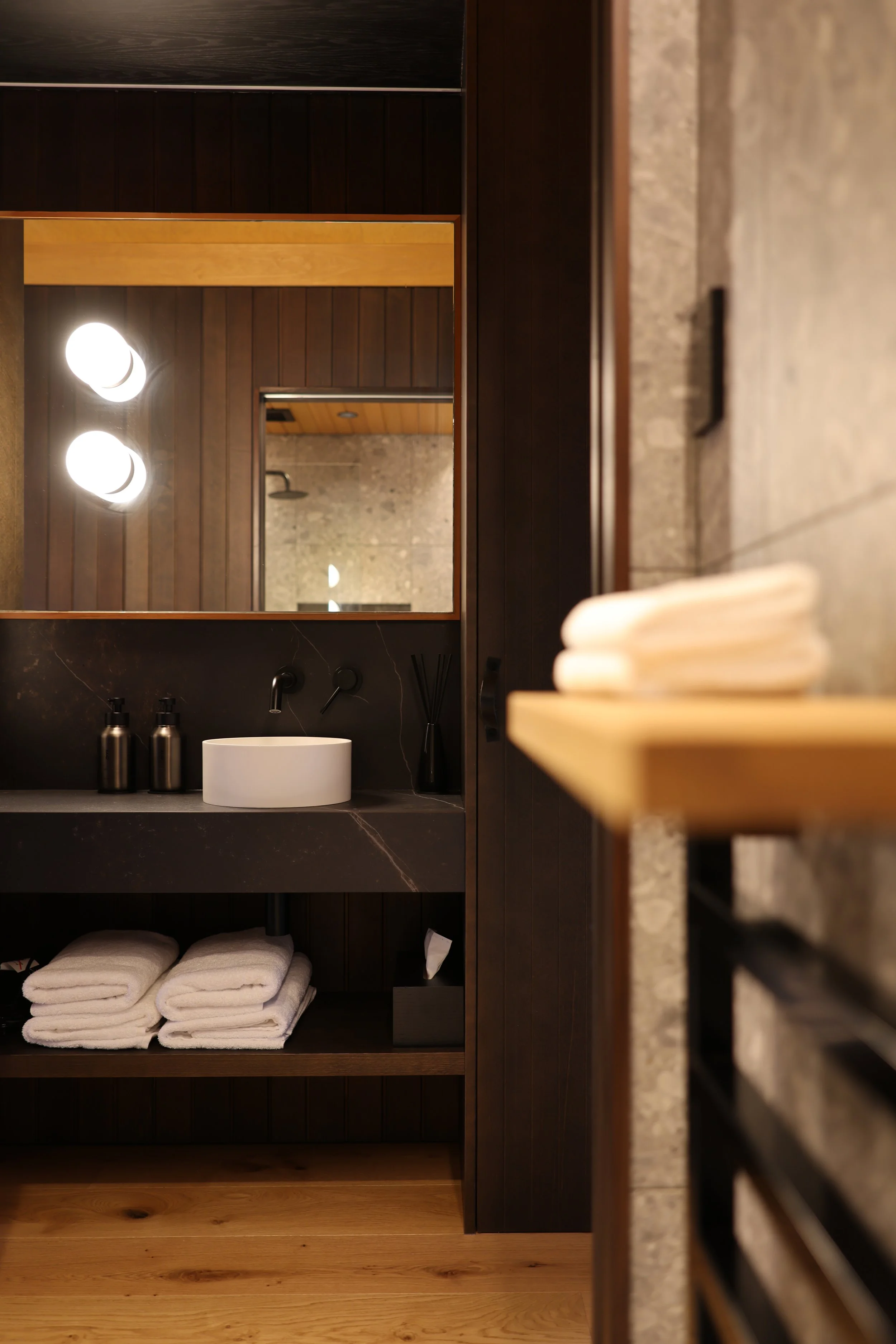 A bathroom with dark wood cabinets, a white round sink, two towels beneath the sink, and shelves with neatly rolled towels. A mirror on the wall reflects two ceiling lights and part of the shower area.