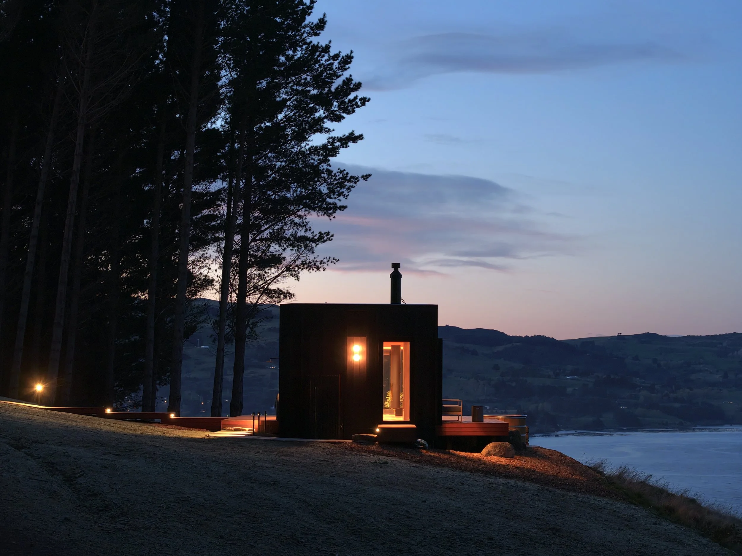 A modern cabin with large glass sliding doors illuminated from inside, and an outdoor hot tub, surrounded by a forest, set against a sunset sky with blue and pink hues. A balcony with outdoor seating is visible.