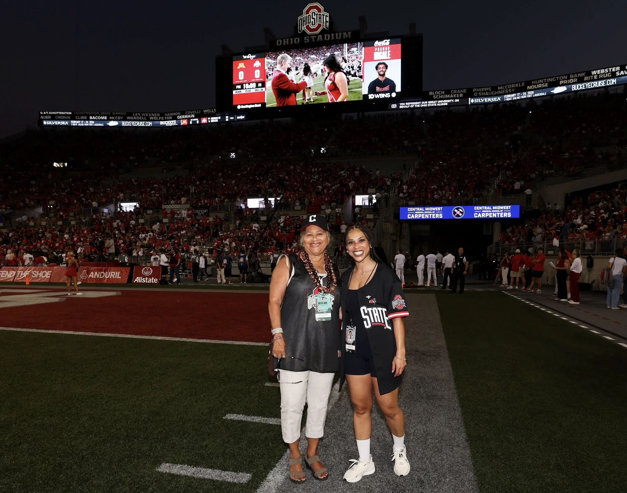 Two women standing on a football field at Ohio Stadium during a nighttime game, wearing Ohio State Buckeyes apparel, smiling at the camera, with a large crowd and scoreboard in the background.